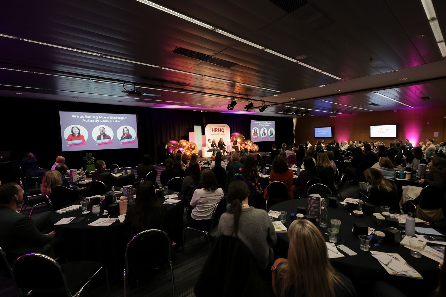 Large conference room filled with attendees seated at tables facing a illuminated stage with speakers and presentation screens.