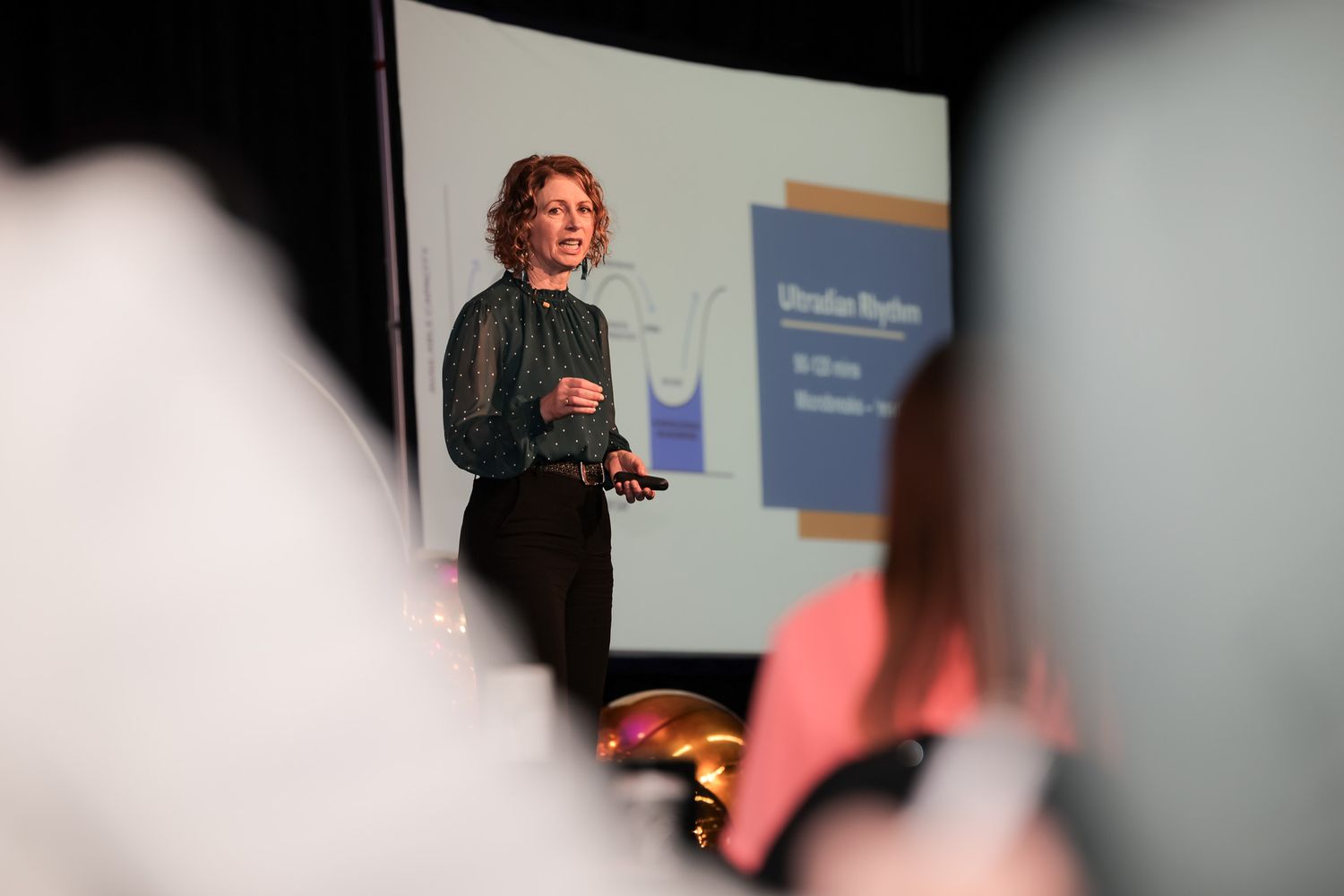 Woman presenting on stage at a conference with a blue projection screen behind her.