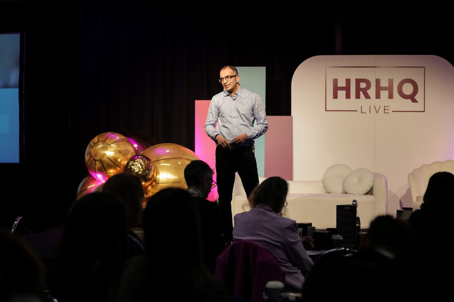 Speaker presenting on stage at HRHQ Live event with golden balloons and audience in foreground.
