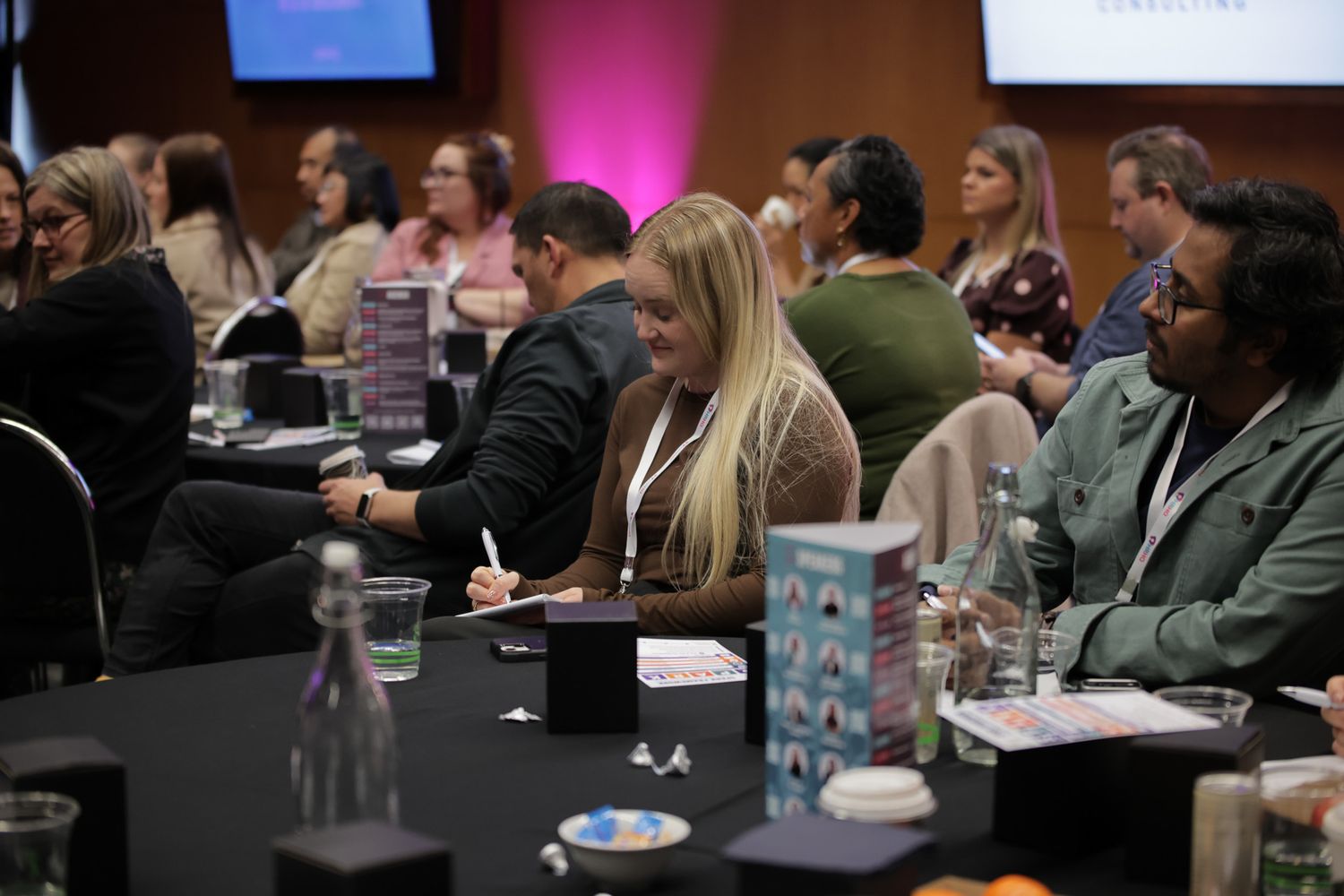 Professionals networking at a conference, seated at tables, taking notes and engaging in discussion in a modern event venue.