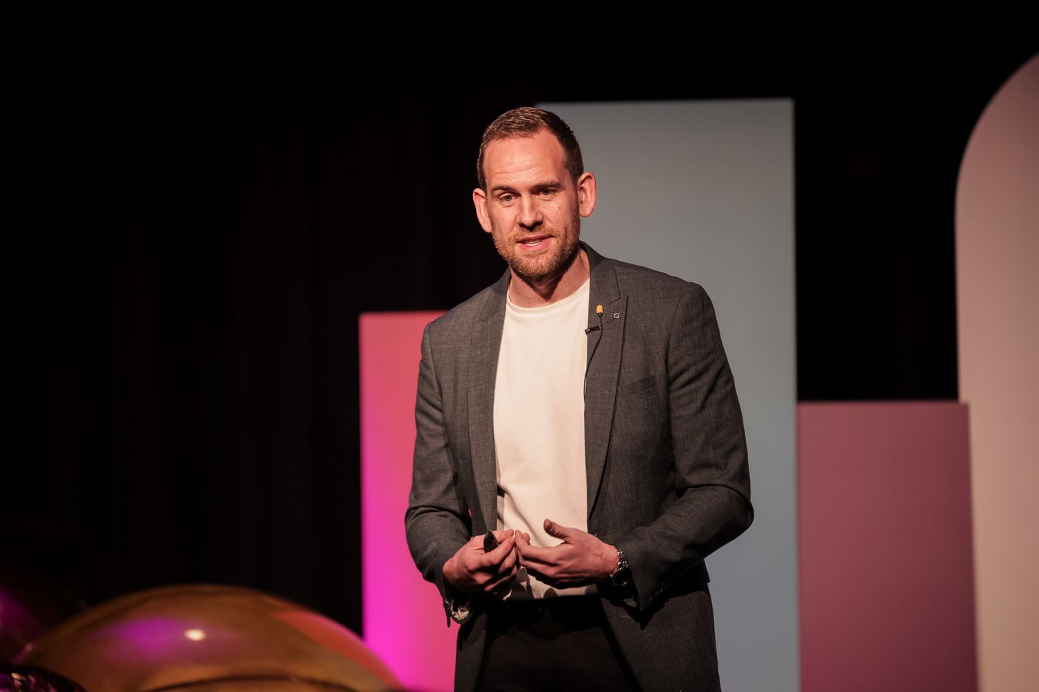 Man in gray blazer speaking on stage with pink and blue backdrop lighting.