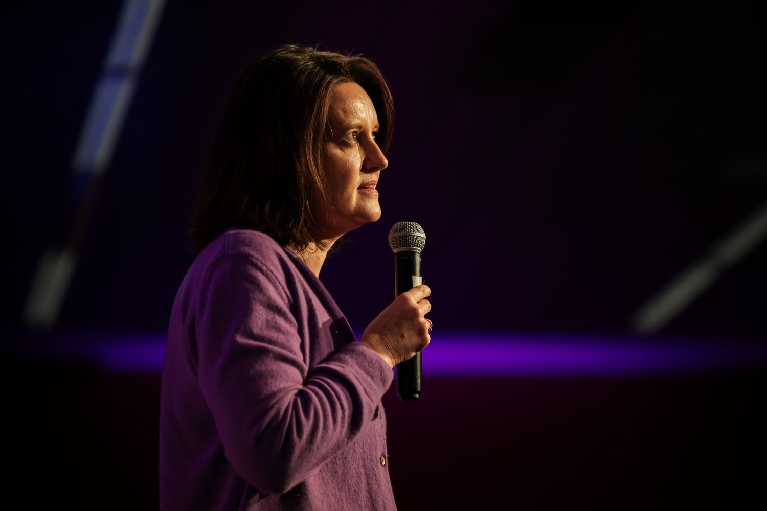 Woman in purple blazer speaking into microphone on stage with colorful lighting backdrop.