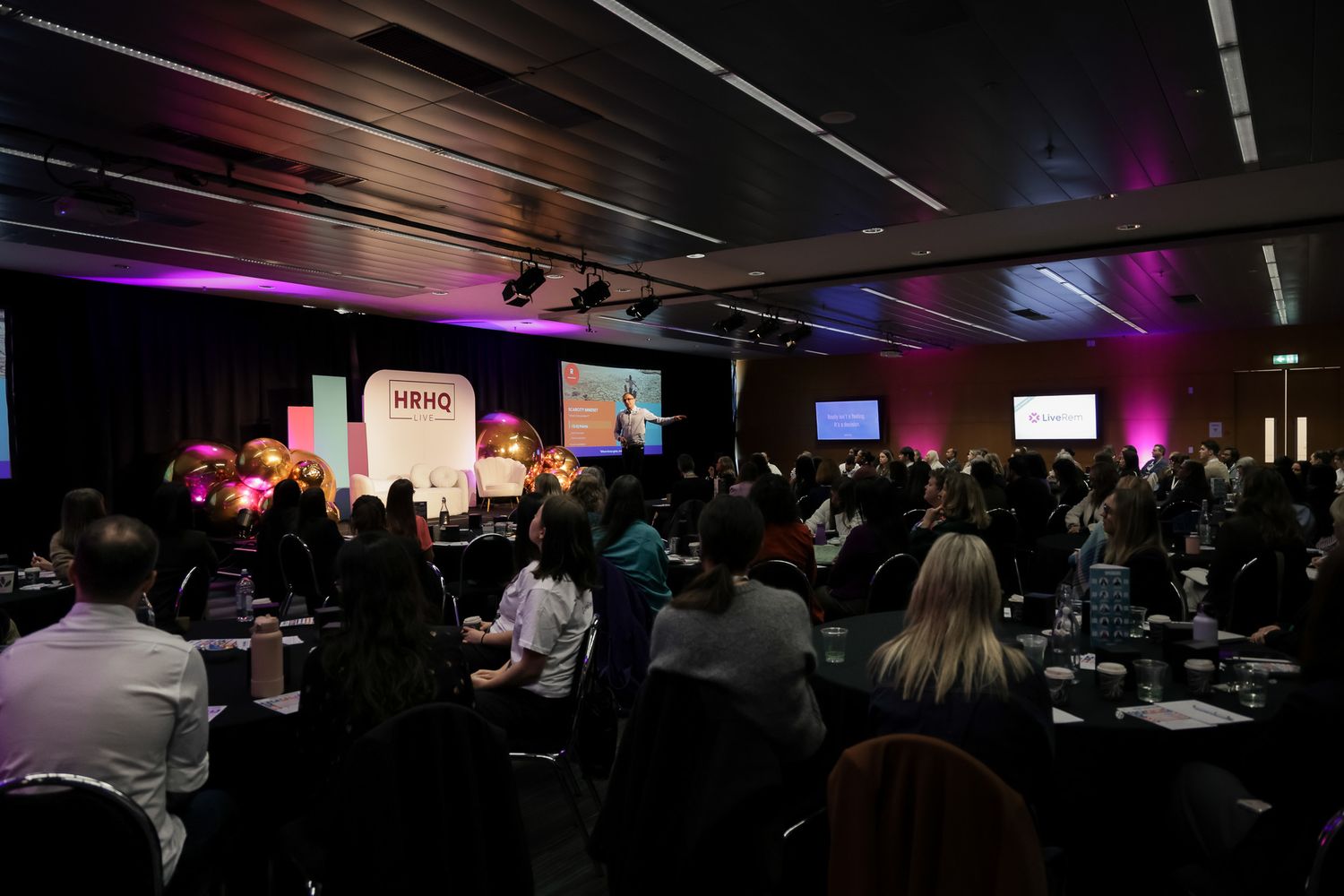 Large conference audience seated at round tables facing a stage with colorful HBHQ branding and presentation screens.