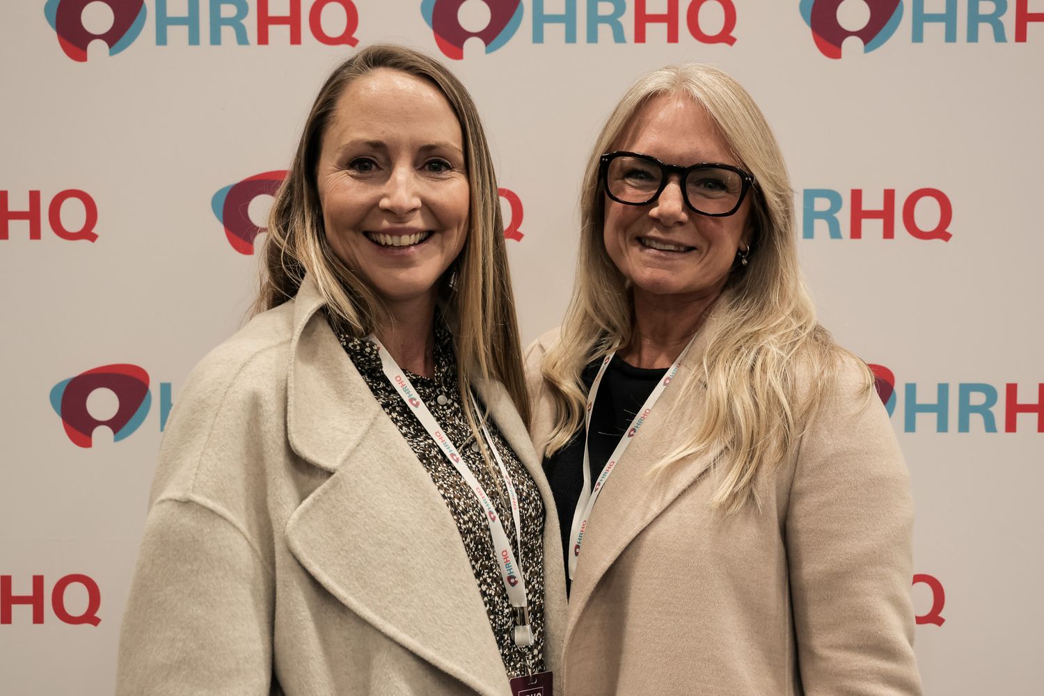 Two women smiling together at an HRHQ event, wearing beige coats in front of a branded backdrop.