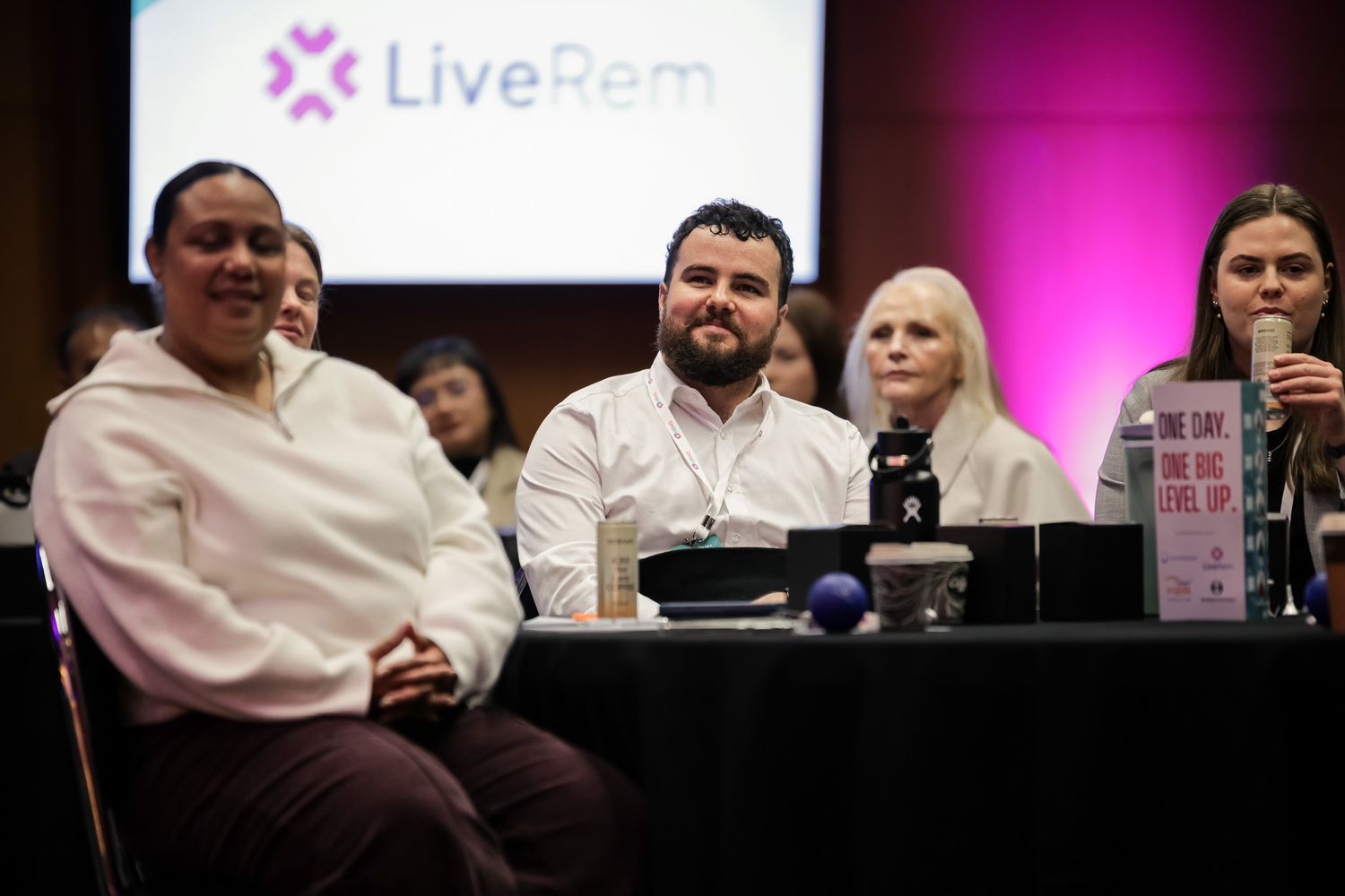 Attendees smile at a LiveRem conference event, seated at a table with a pink branded backdrop.