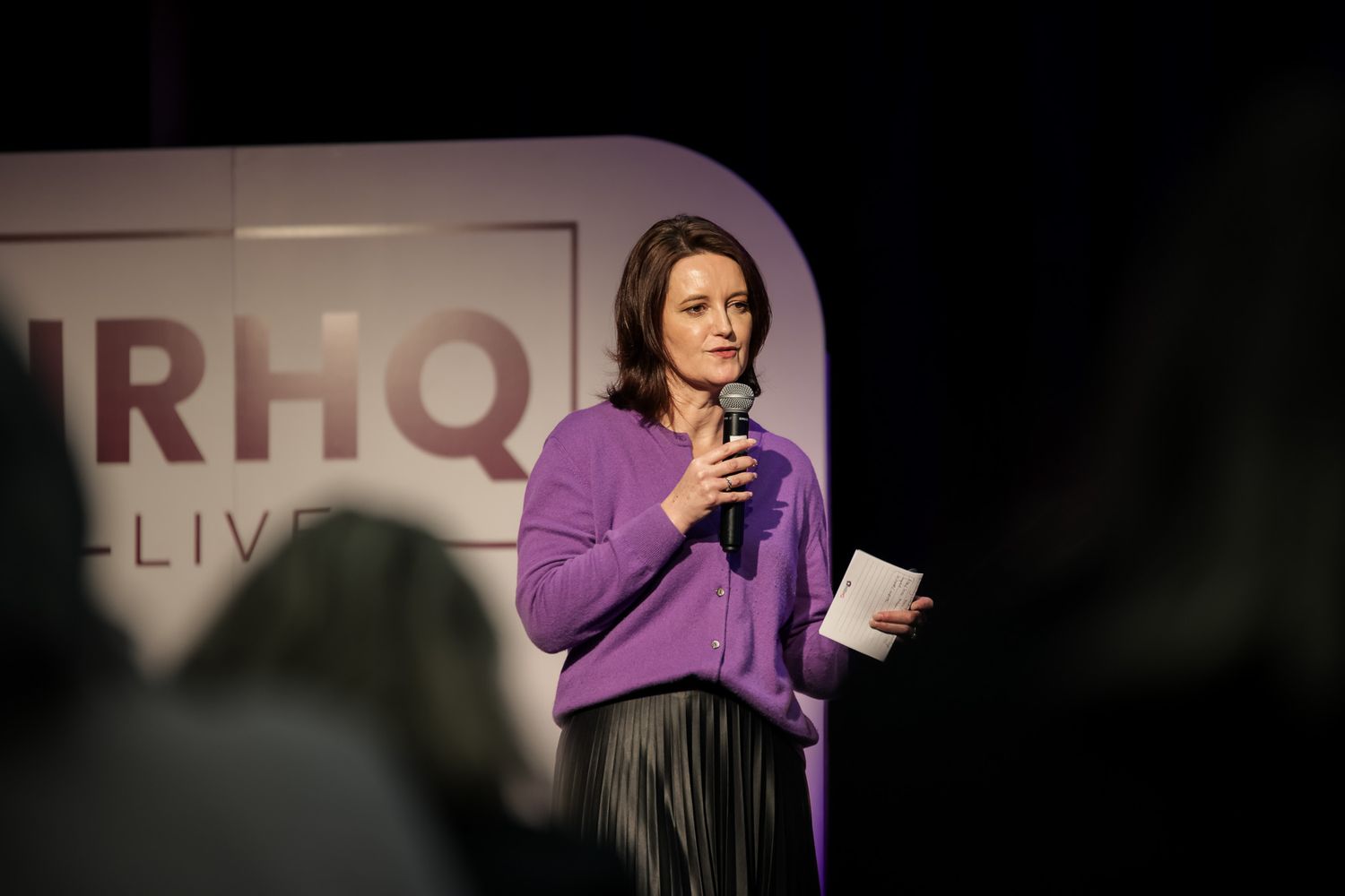 Woman in purple cardigan speaks at HRHQ Live event, holding microphone and notes on stage.