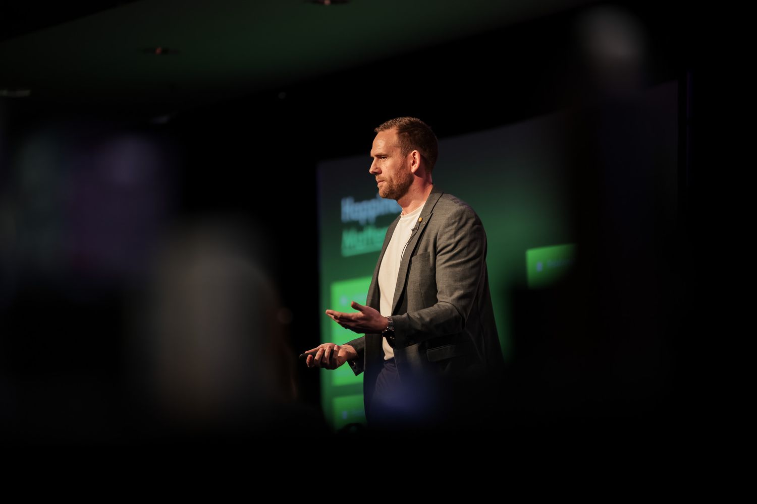 A male speaker presents on stage at a conference, illuminated by green lighting with a presentation screen behind him.