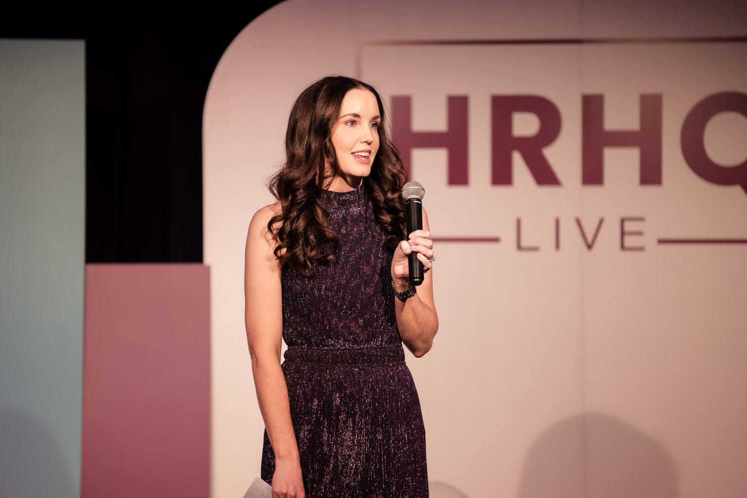 Woman in purple dress speaking on stage at HRHC Live event conference.