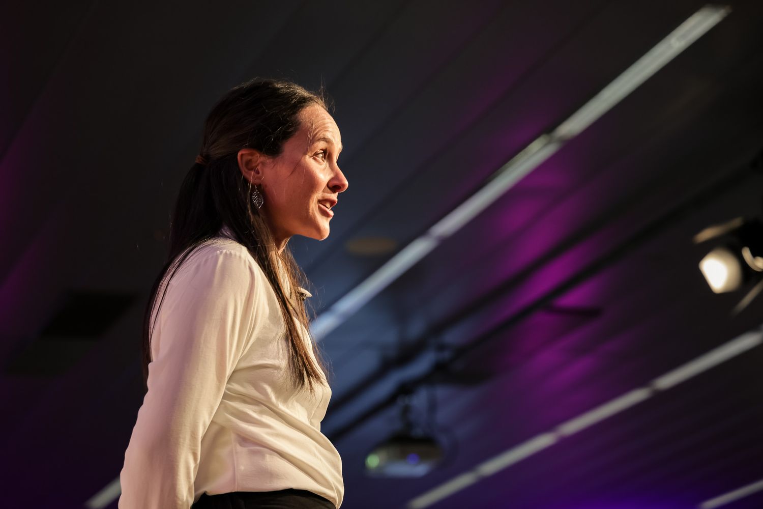 Woman in white blouse speaking on stage with purple lighting backdrop at a professional event.