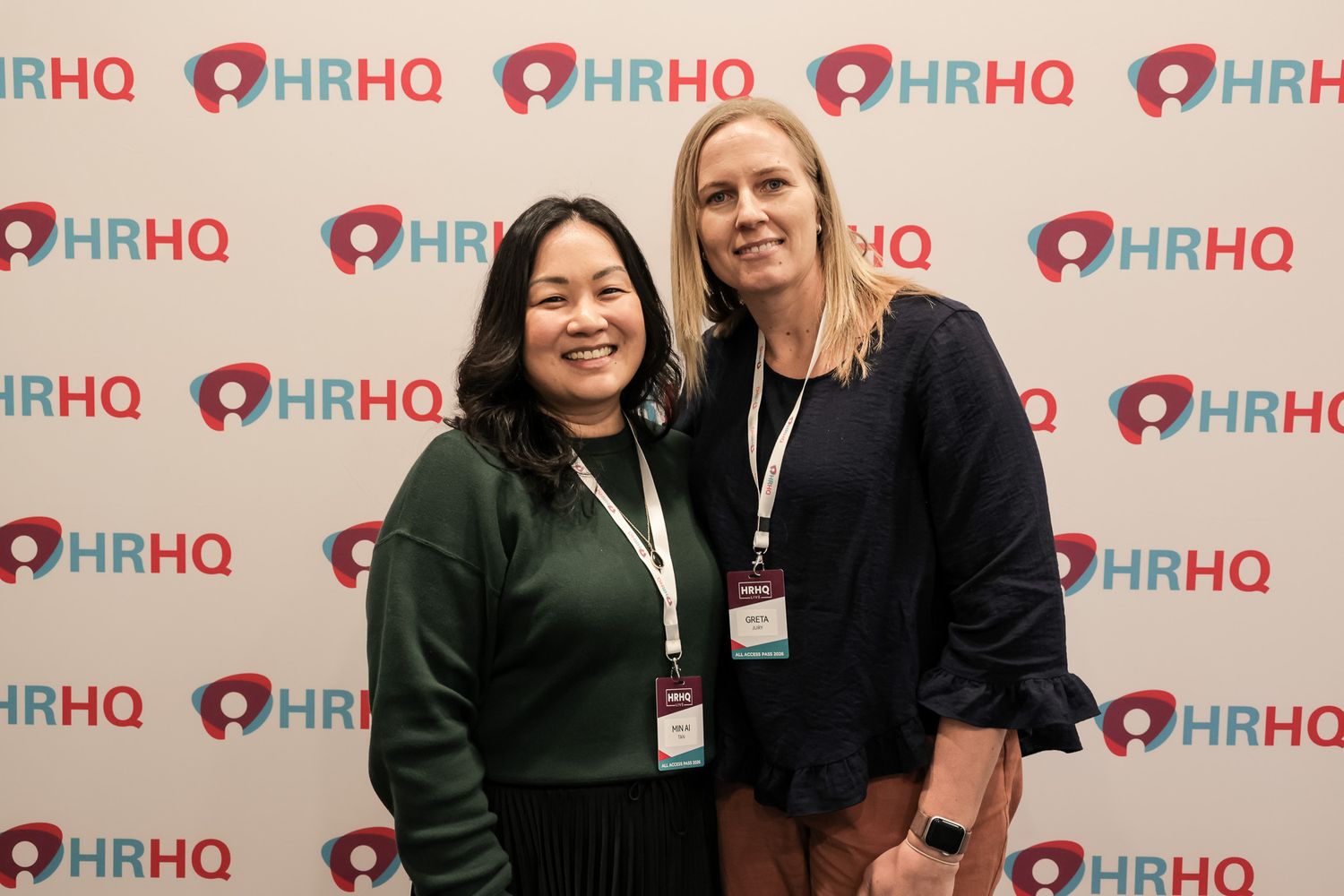 Two women smiling at an HRHQ conference event, posing in front of a branded step-and-repeat backdrop.