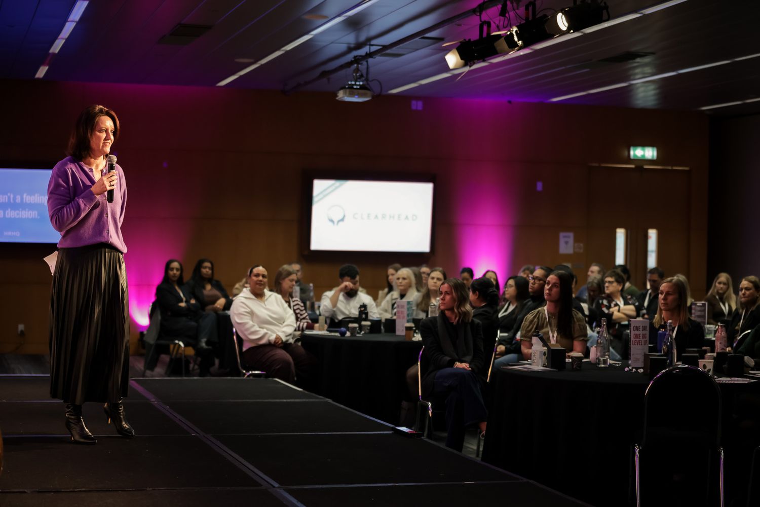 Woman speaker addresses audience at a corporate event with pink lighting in a modern conference hall.