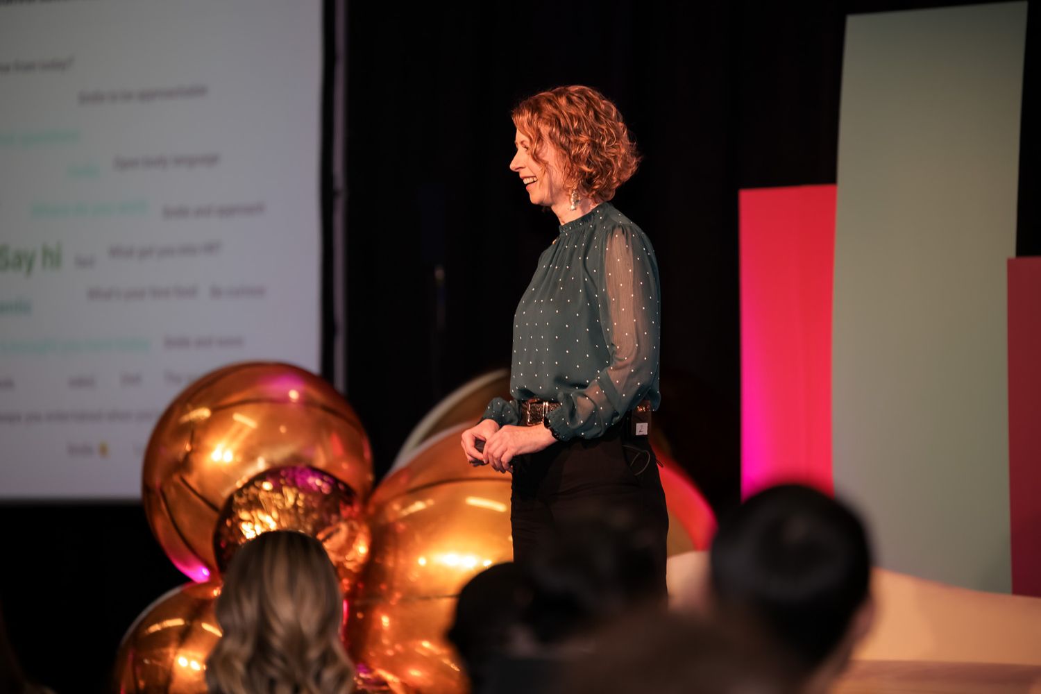 Woman with curly hair presenting on stage at a conference with gold balloons and a projection screen behind her.
