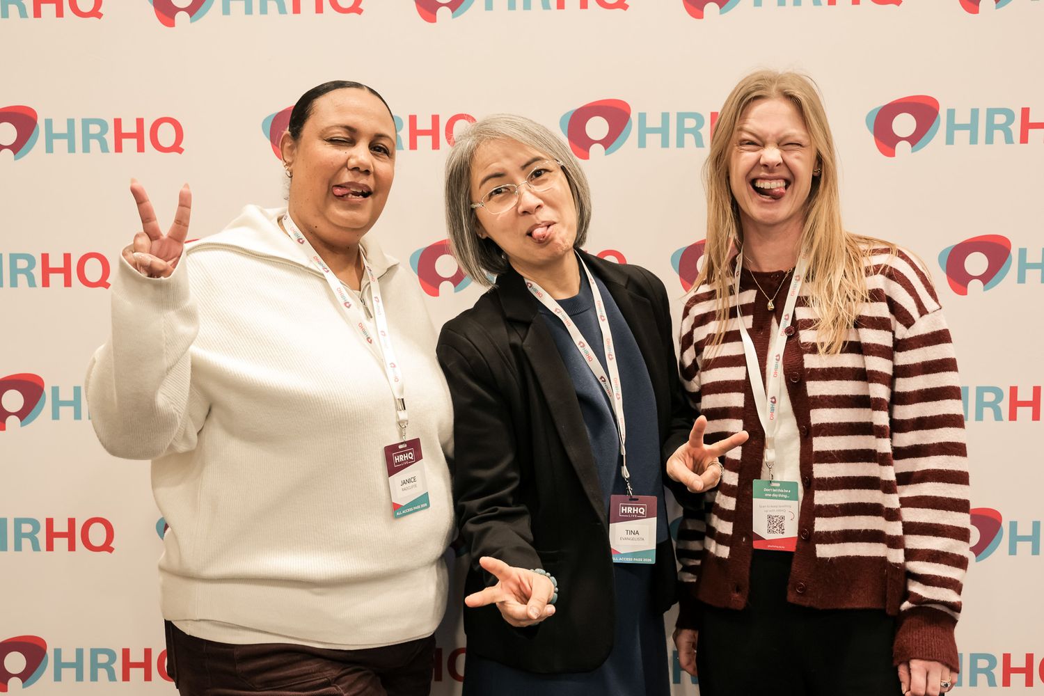 Three women posing and smiling at an HRHQ conference event backdrop, making fun gestures for the camera.