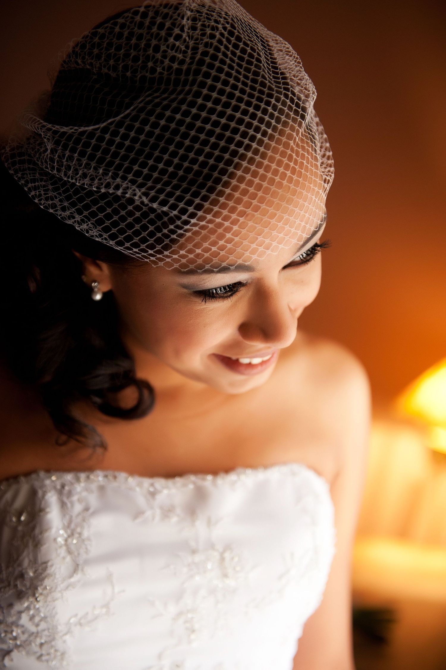 Close-up details of a white lace wedding dress and birdcage veil in warm lighting.
