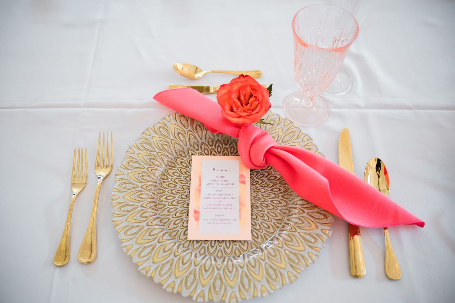 Place setting with coral napkin, gold charger plate, and orange rose on white tablecloth.