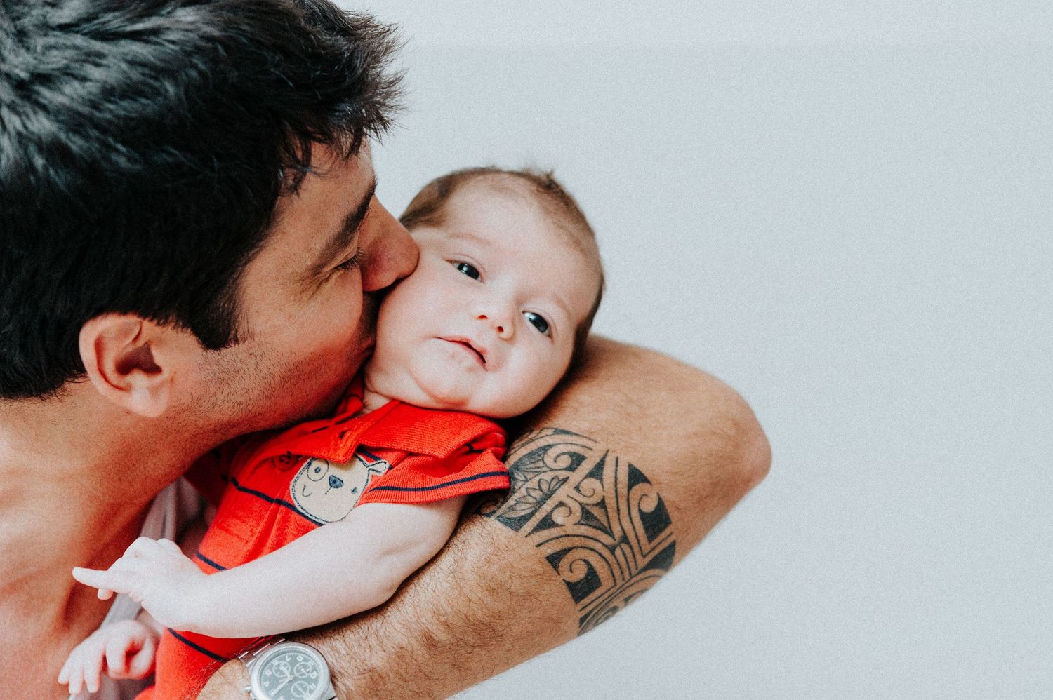 Baby in red outfit being held and kissed against light background.