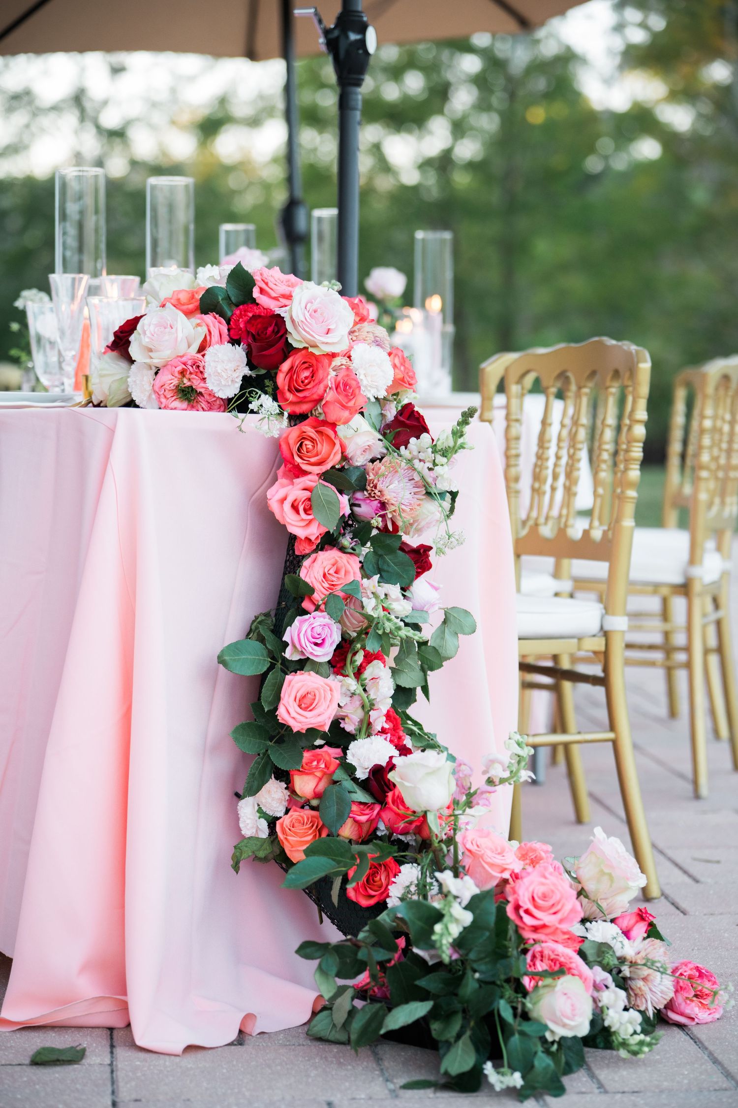 Pink and coral rose floral arrangements cascading down tables with gold chairs at outdoor reception.