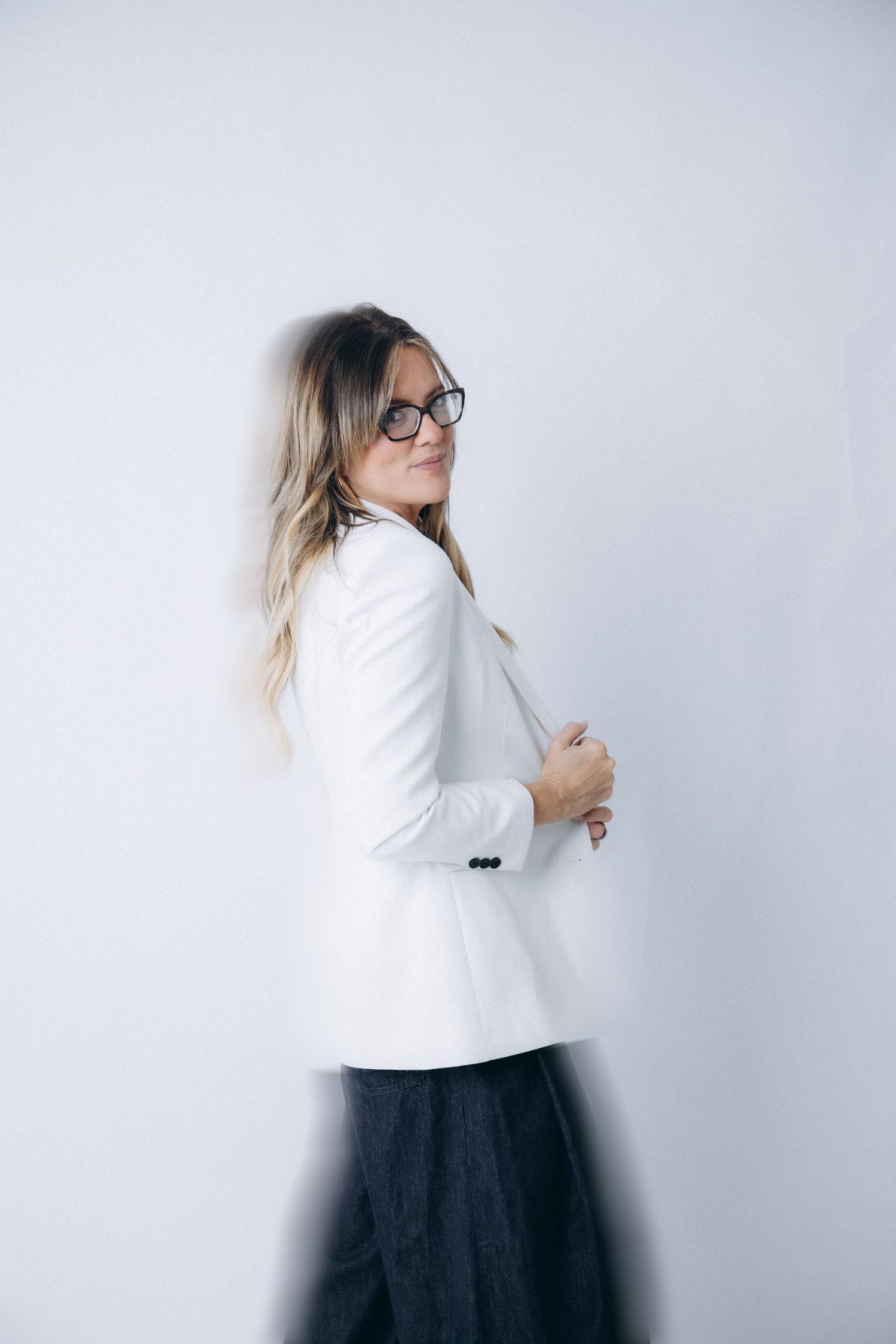 Professional wearing white blazer and glasses poses playfully against white background.