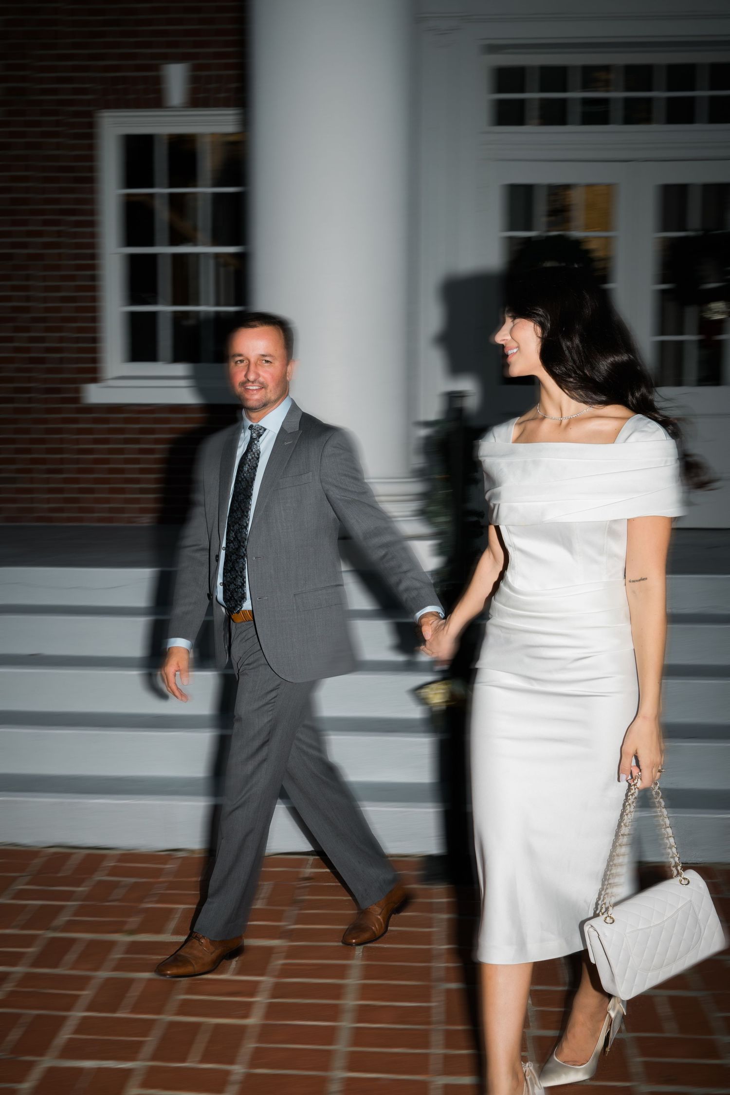 A couple in formal attire walking along a brick pathway at dusk.