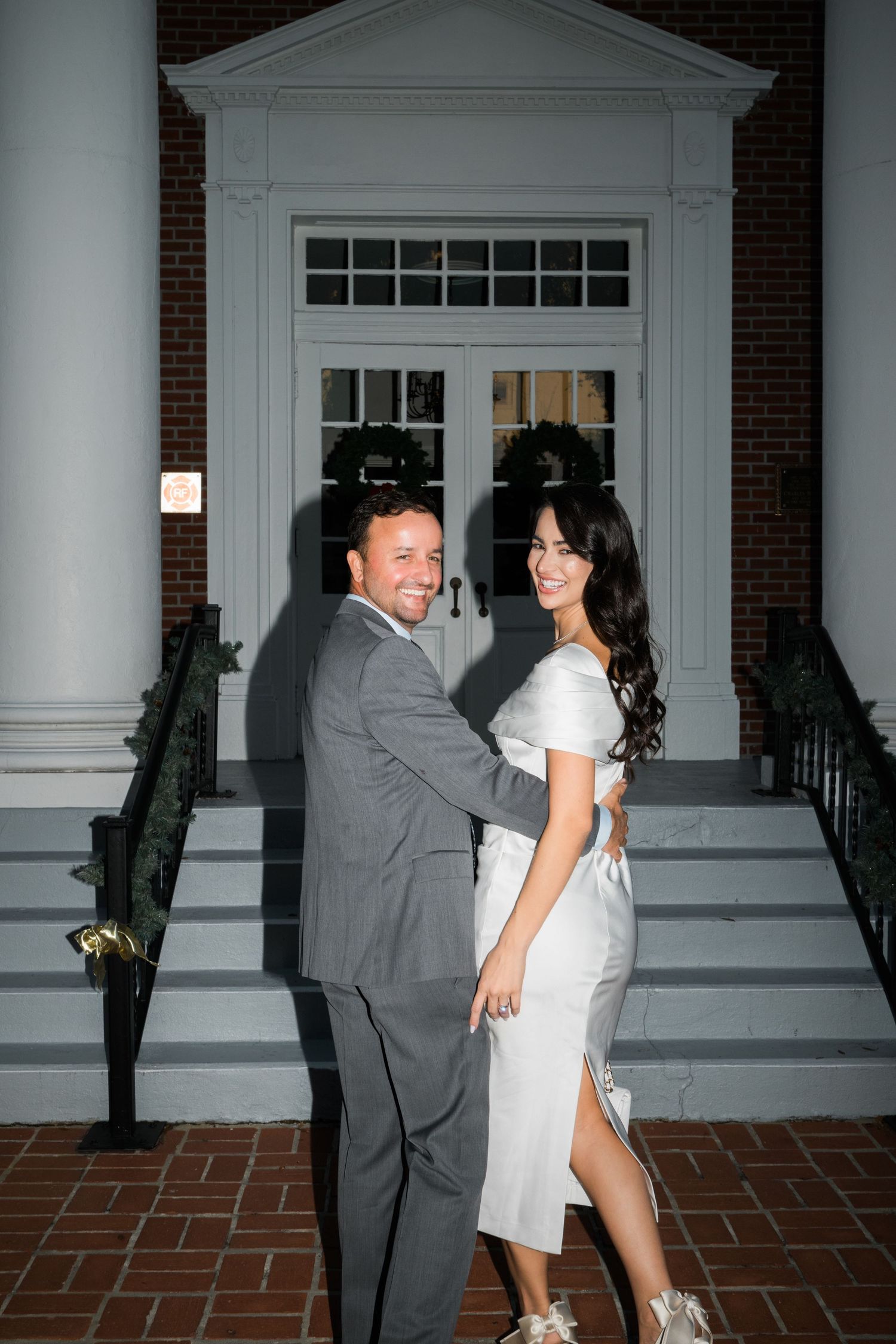 A playful couple poses by an elegant white doorway of a historic building at night.