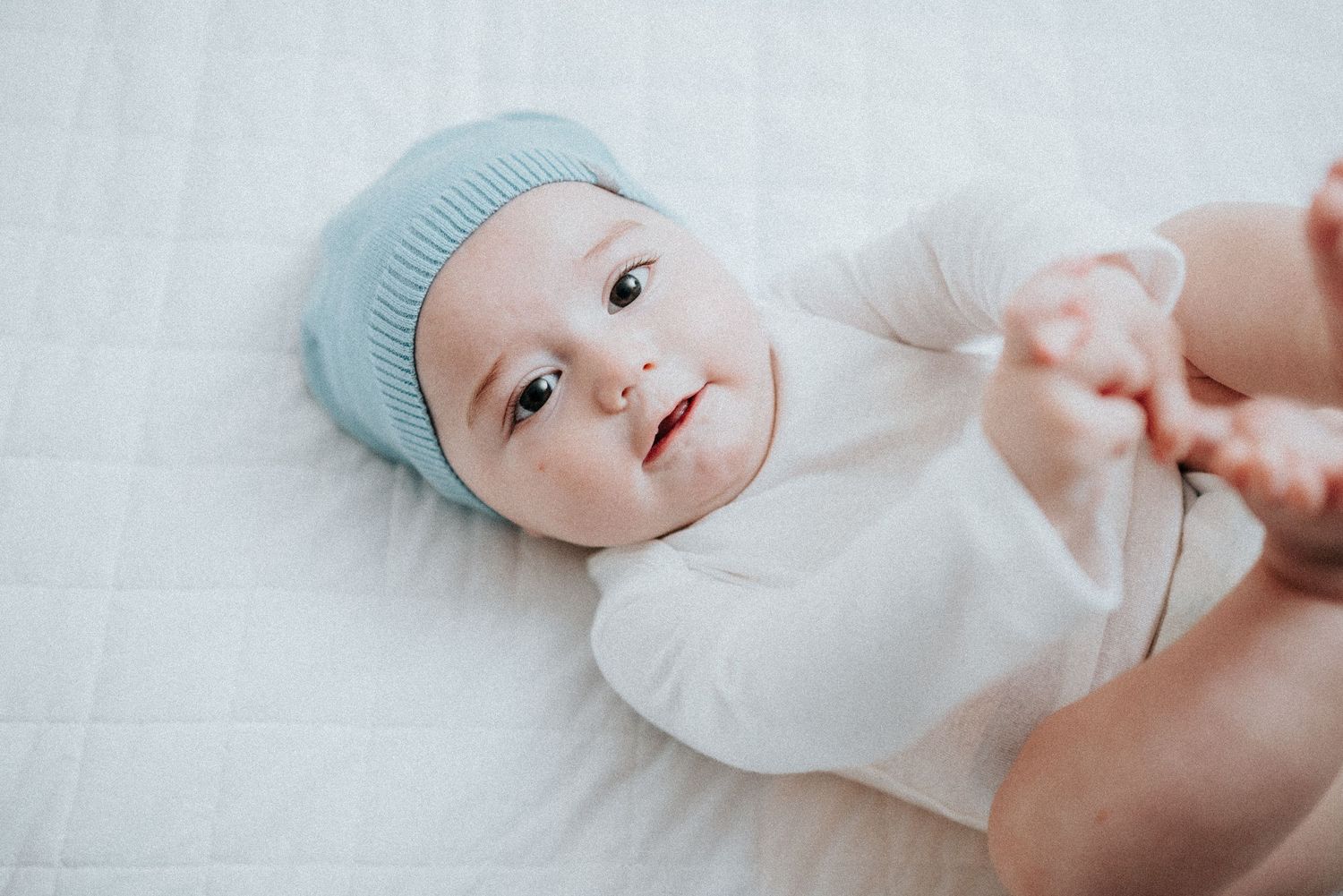 Baby in blue hat and white onesie rests on light colored bedding.