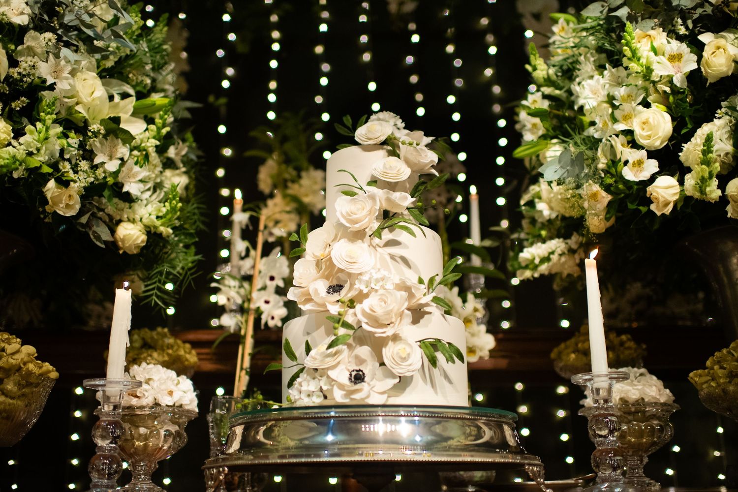 Elegant white wedding cake decorated with floral designs and surrounded by string lights.