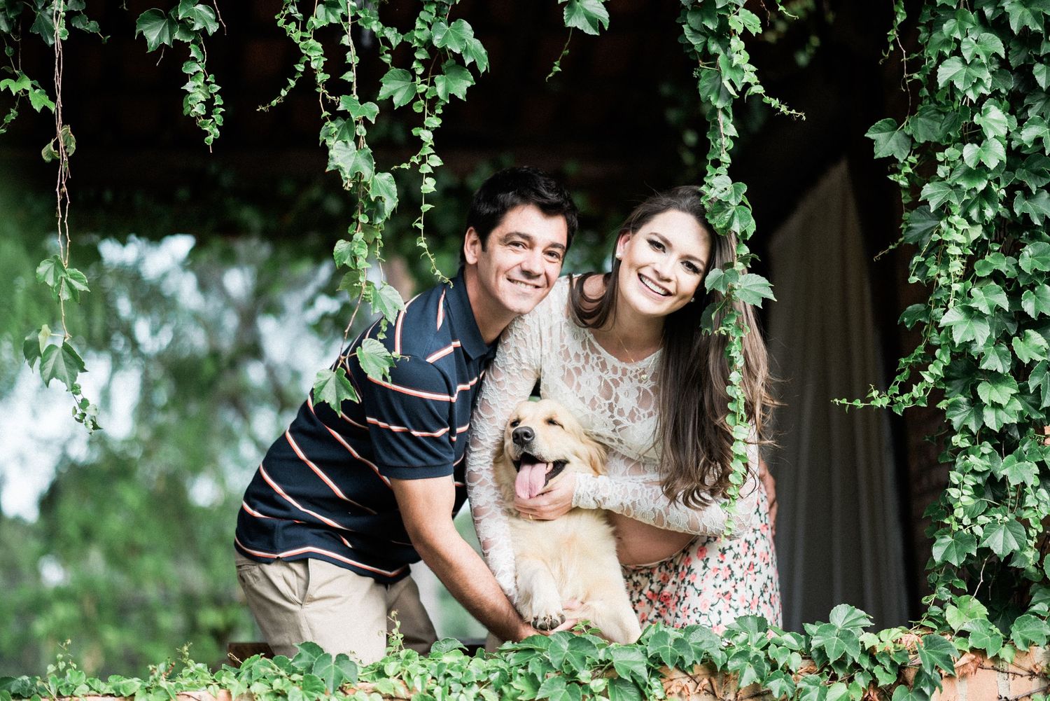 Couple with their dog posing together under lush green trees.