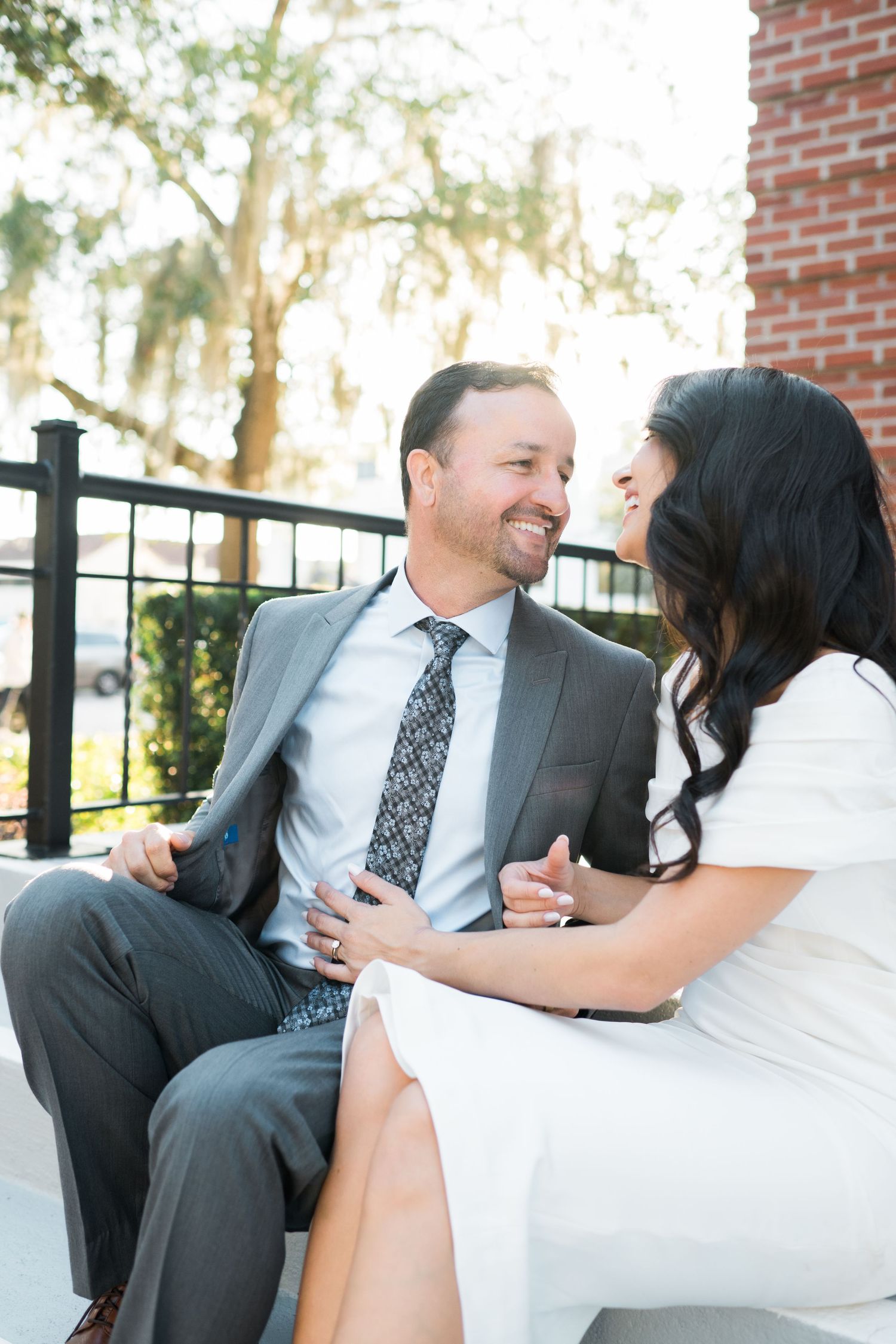 Happy couple in formal attire sharing laughter while sitting outdoors.