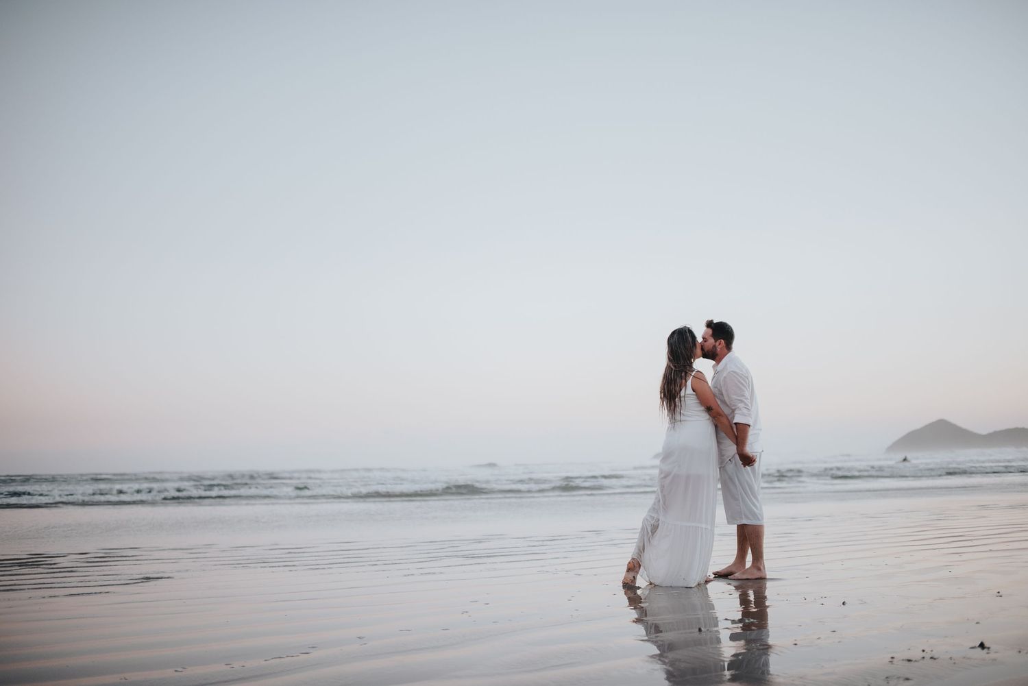 Romantic beach photoshoot with couple in white clothing against misty ocean backdrop.