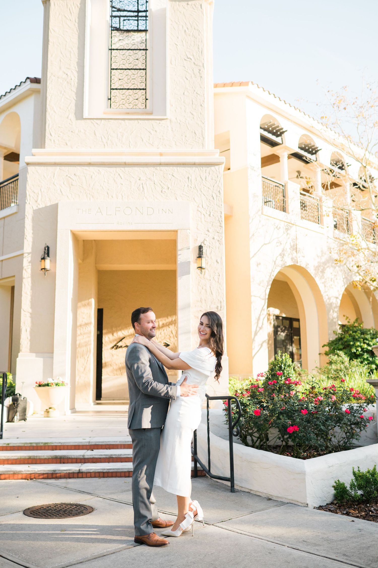 A couple shares a romantic moment in front of a Mediterranean-style building with white walls and red flowers.