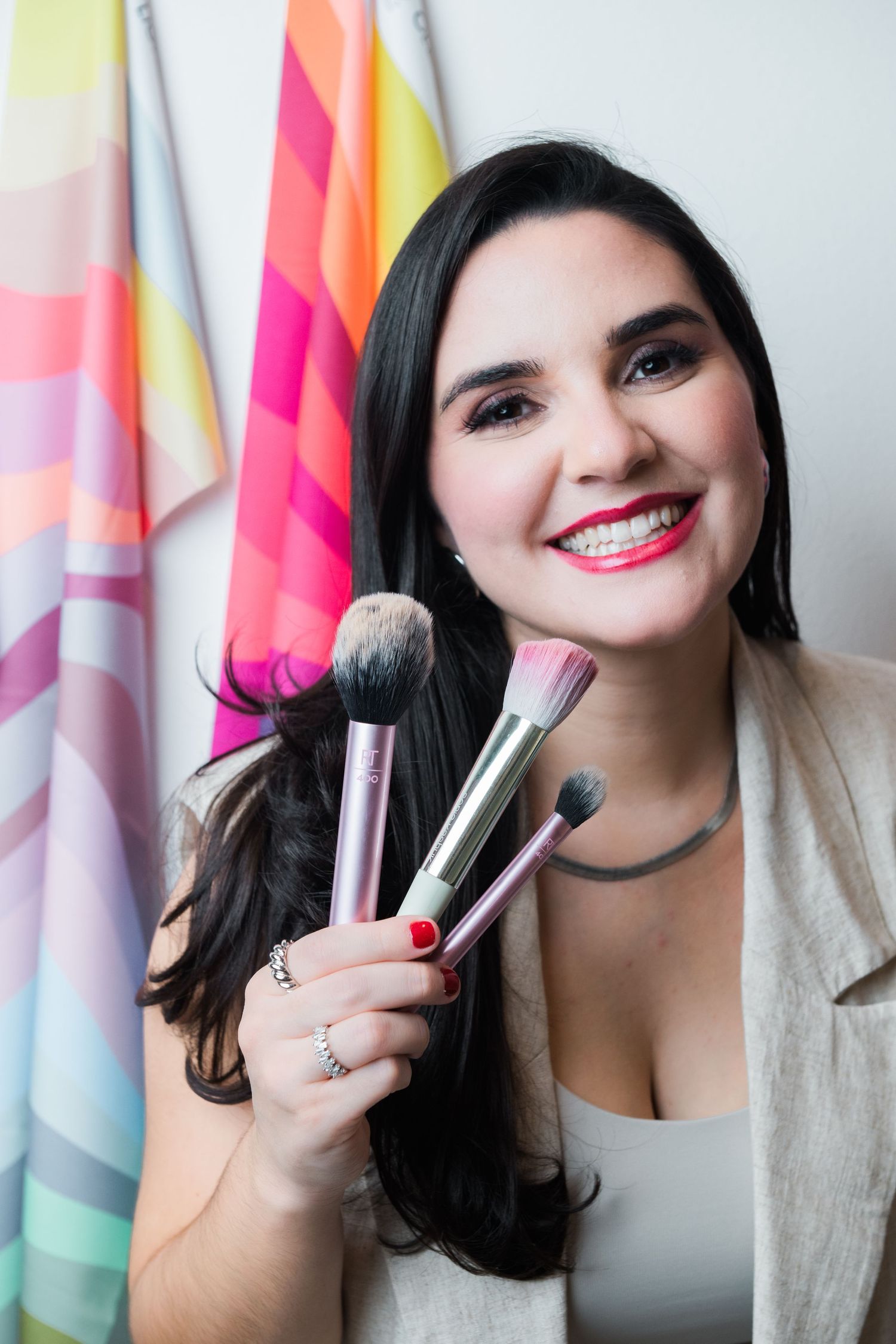 Beauty influencer holding makeup brushes smiles while posing against colorful striped background.