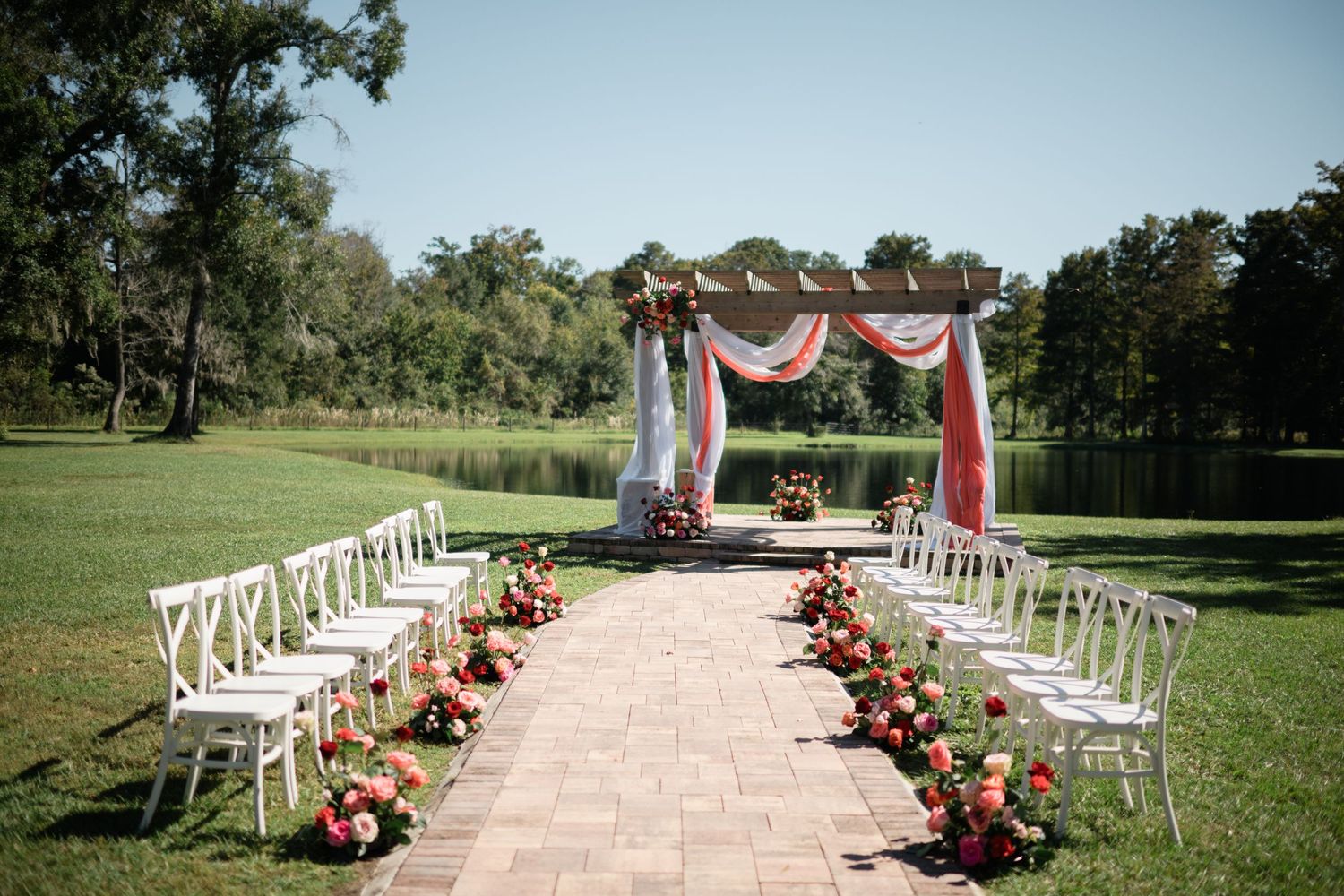 White folding chairs line a stone pathway leading to a wooden wedding arch decorated with coral draping.