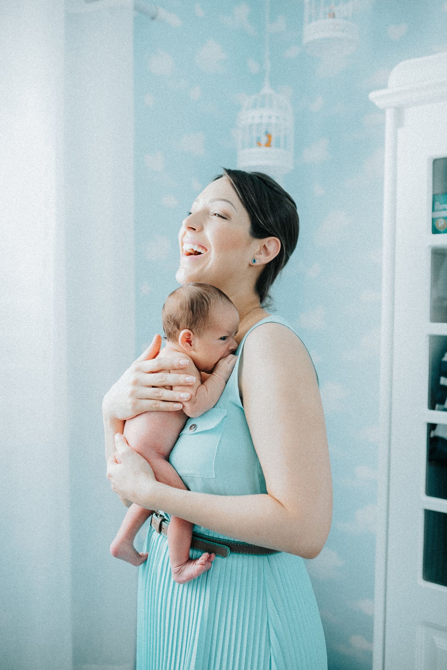 Mother in turquoise dress holding newborn baby against blue wallpapered room.