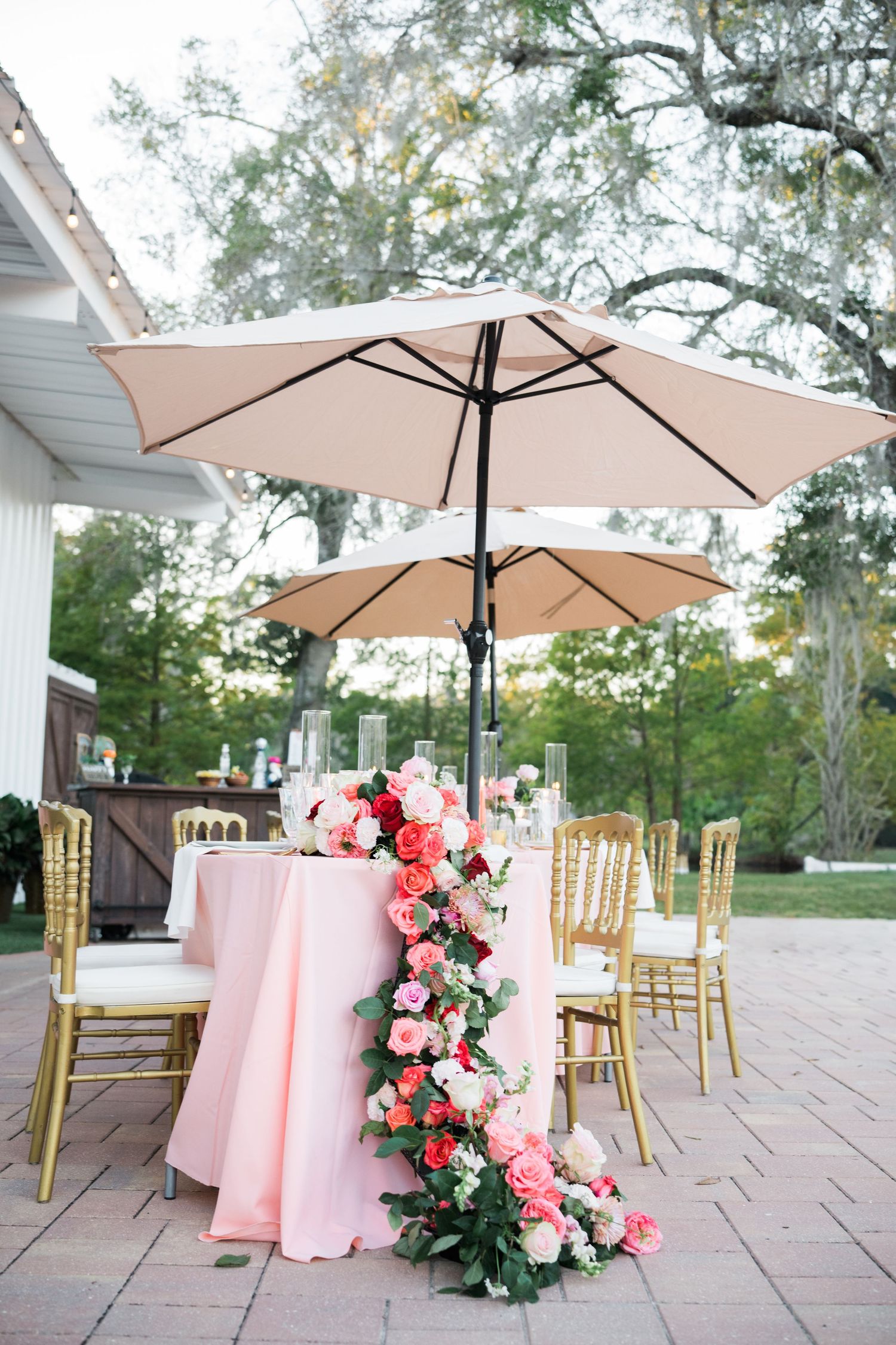 Garden wedding reception setup featuring pink tablecloths, gold chairs, and floral arrangements under umbrellas.
