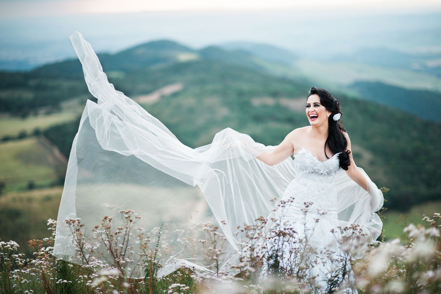 White wedding dress flowing in mountain breeze against scenic landscape backdrop.