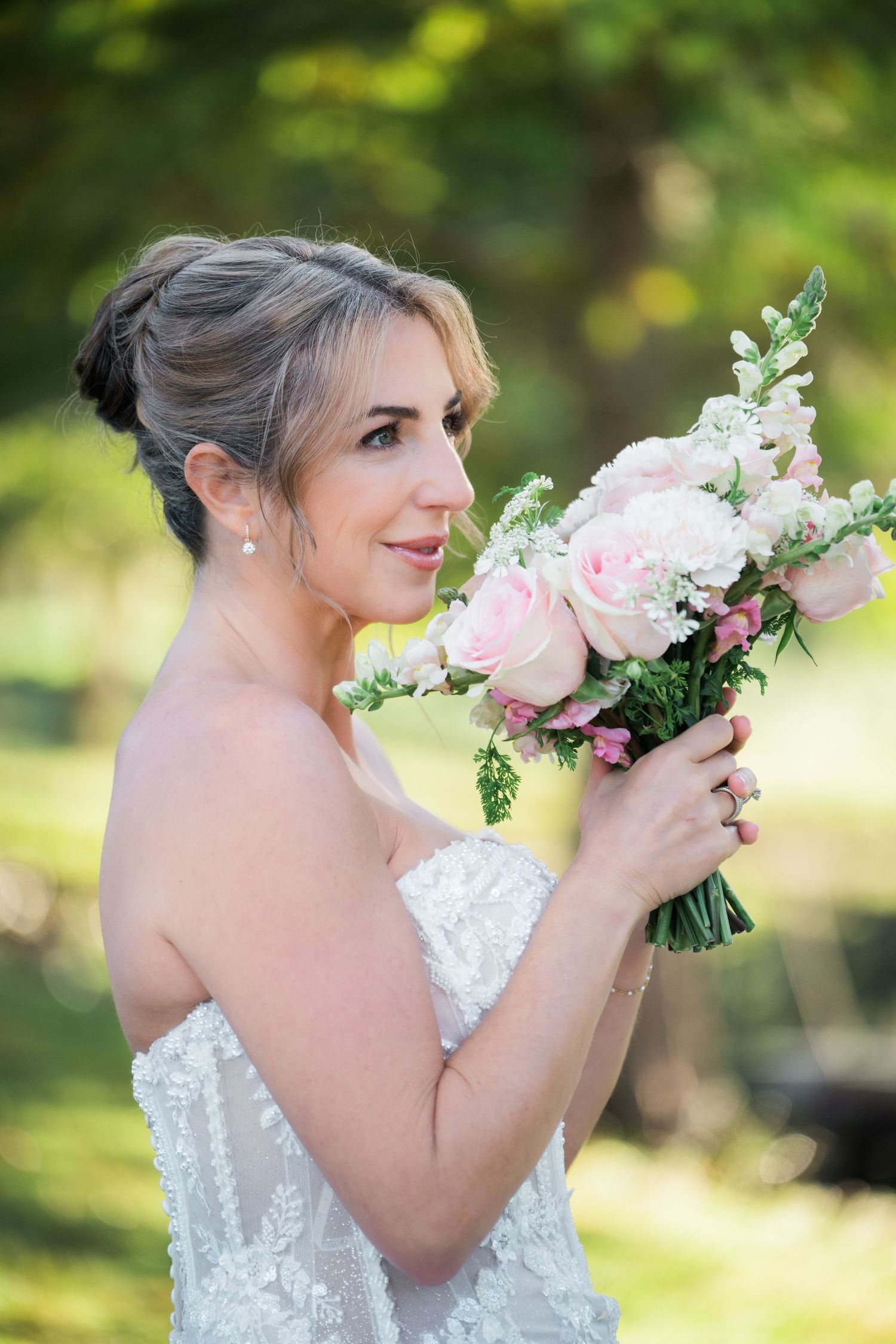 Bride in white lace dress holds pink and white floral bouquet in garden setting.