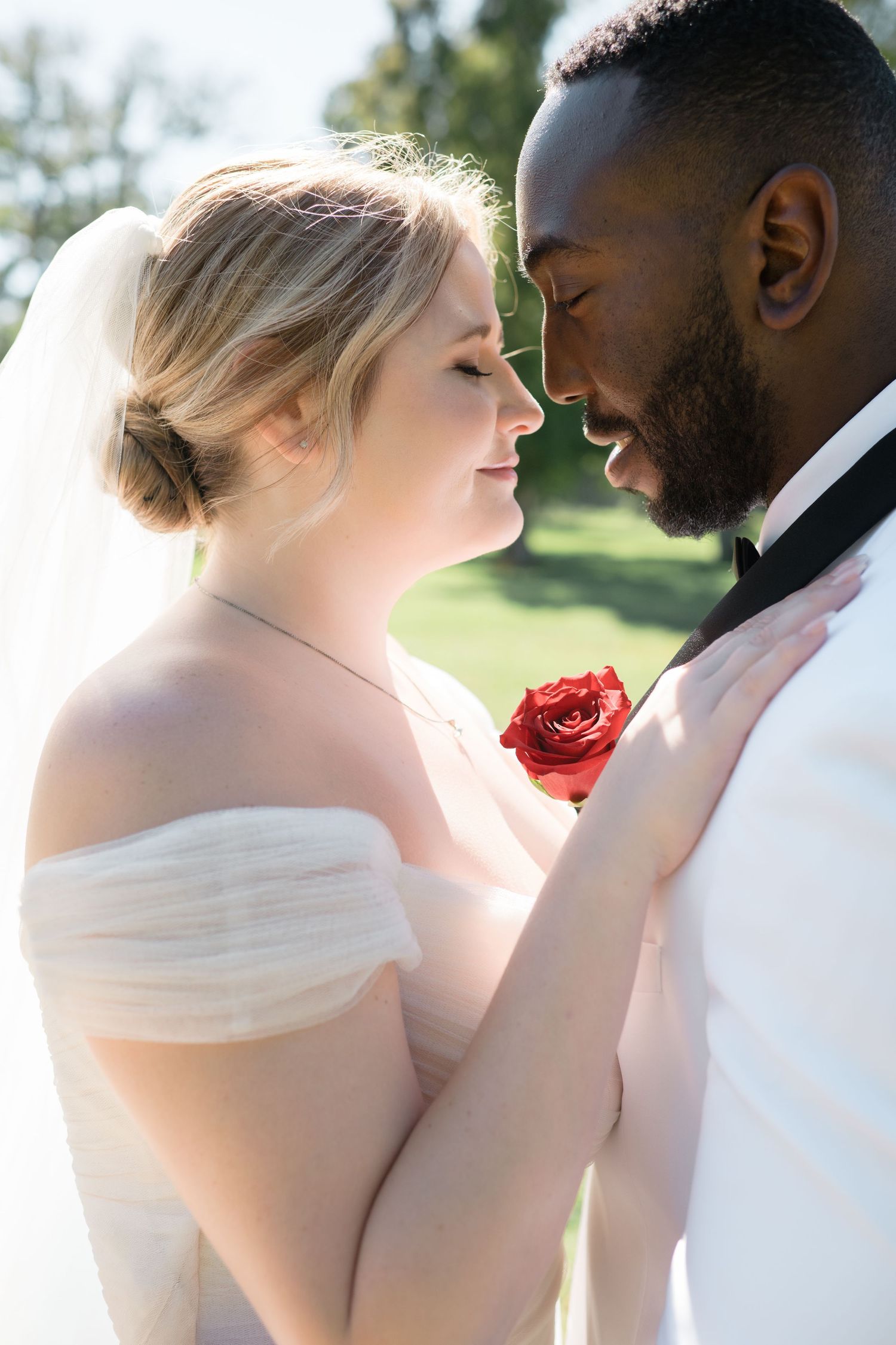 Intimate wedding portrait captured in natural sunlight with red rose accent.