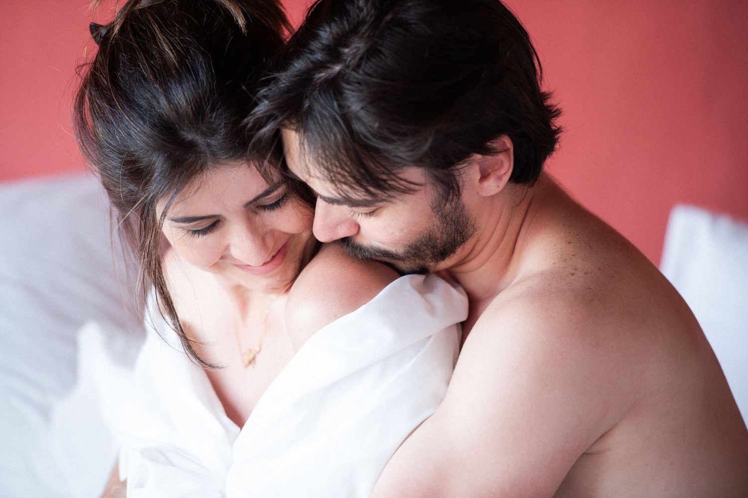 Couple sharing intimate moments on white bedding against red wall background.