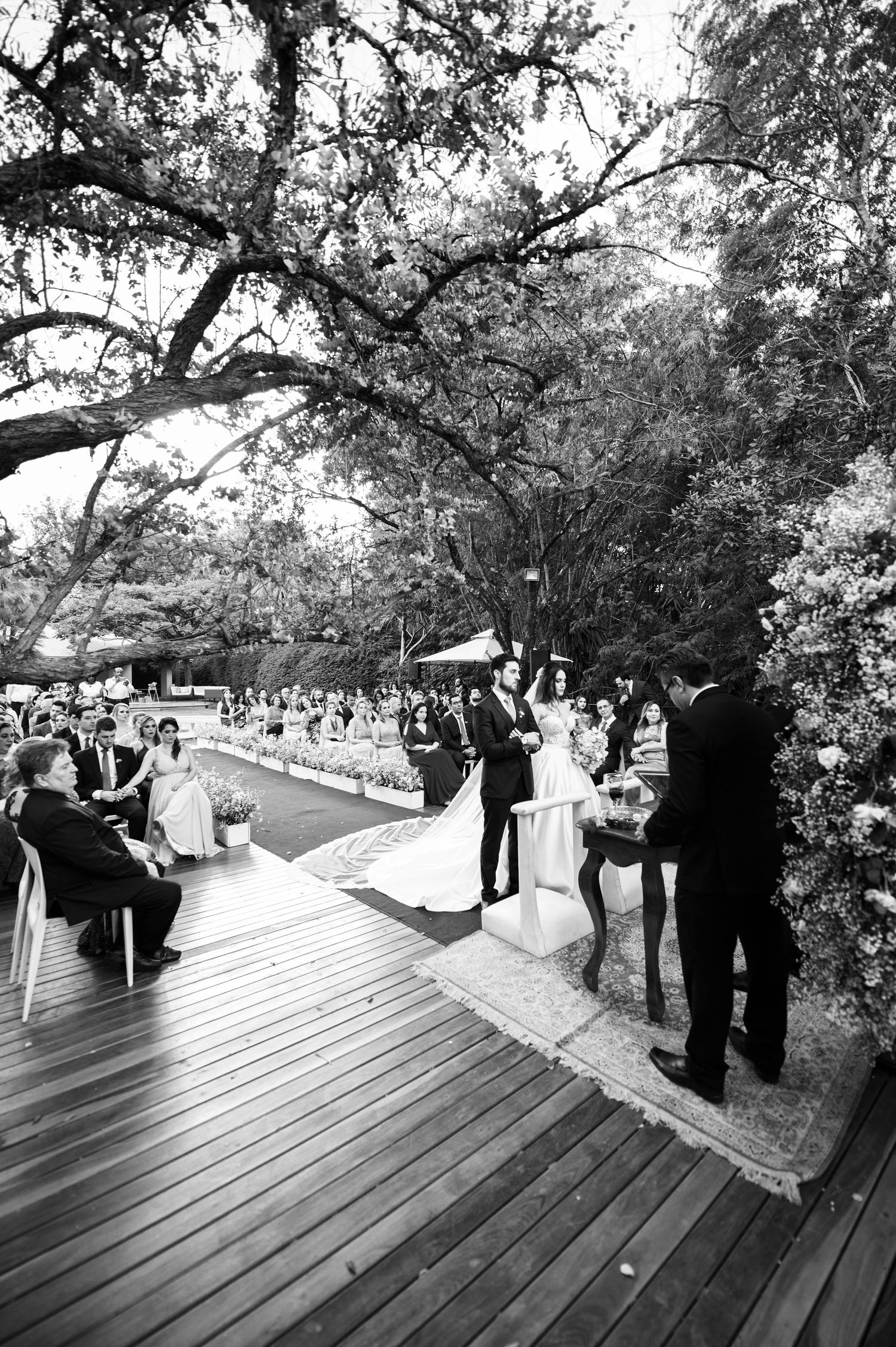 Black and white panorama of wedding guests gathering on a wooden deck under trees.