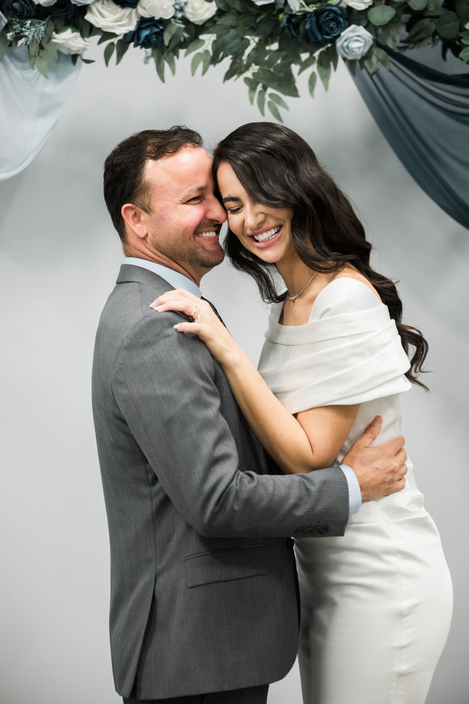 Couple embraces and laughs together against grey wall with floral decorations.