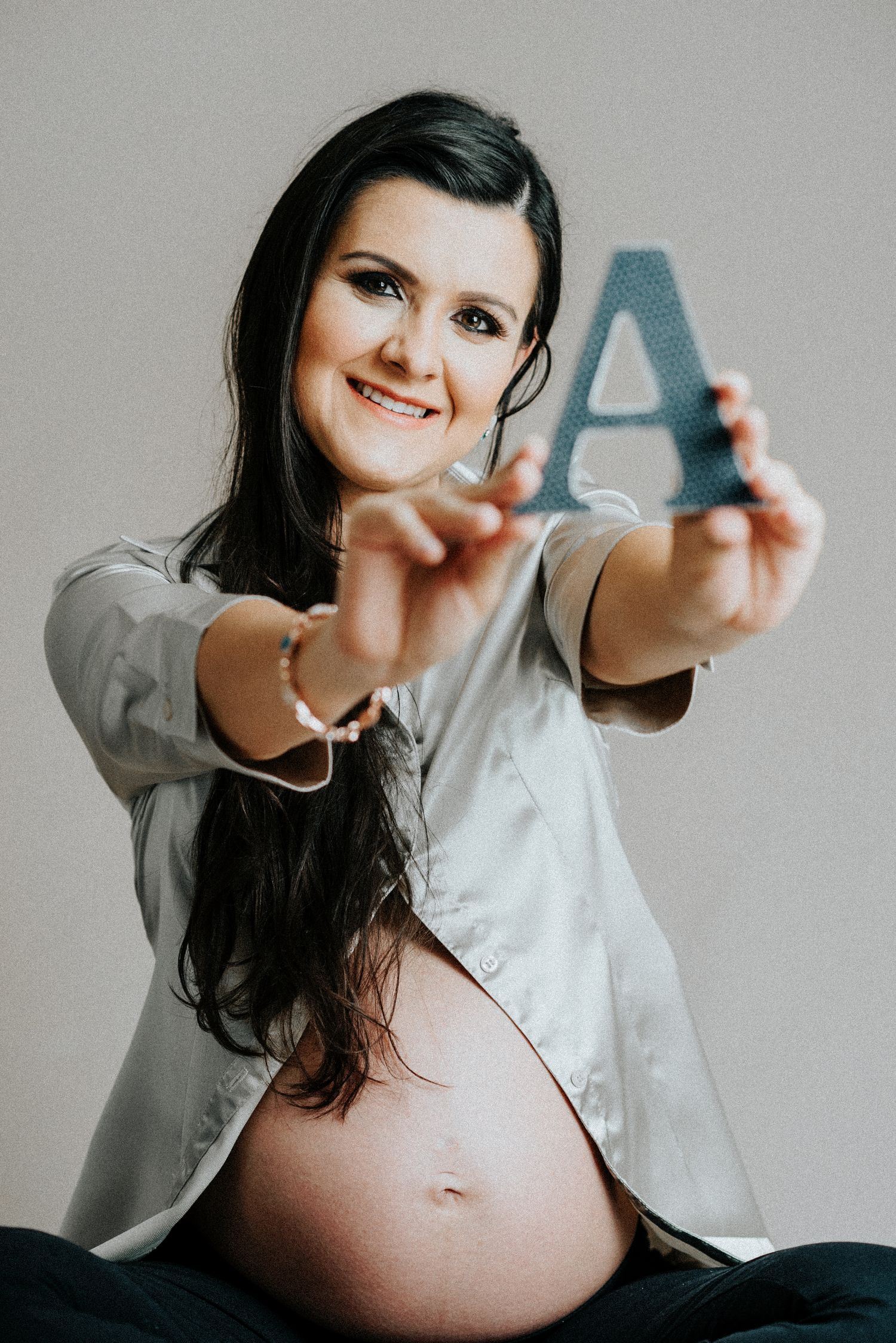 Expectant woman in white shirt holds up baby items against grey background.