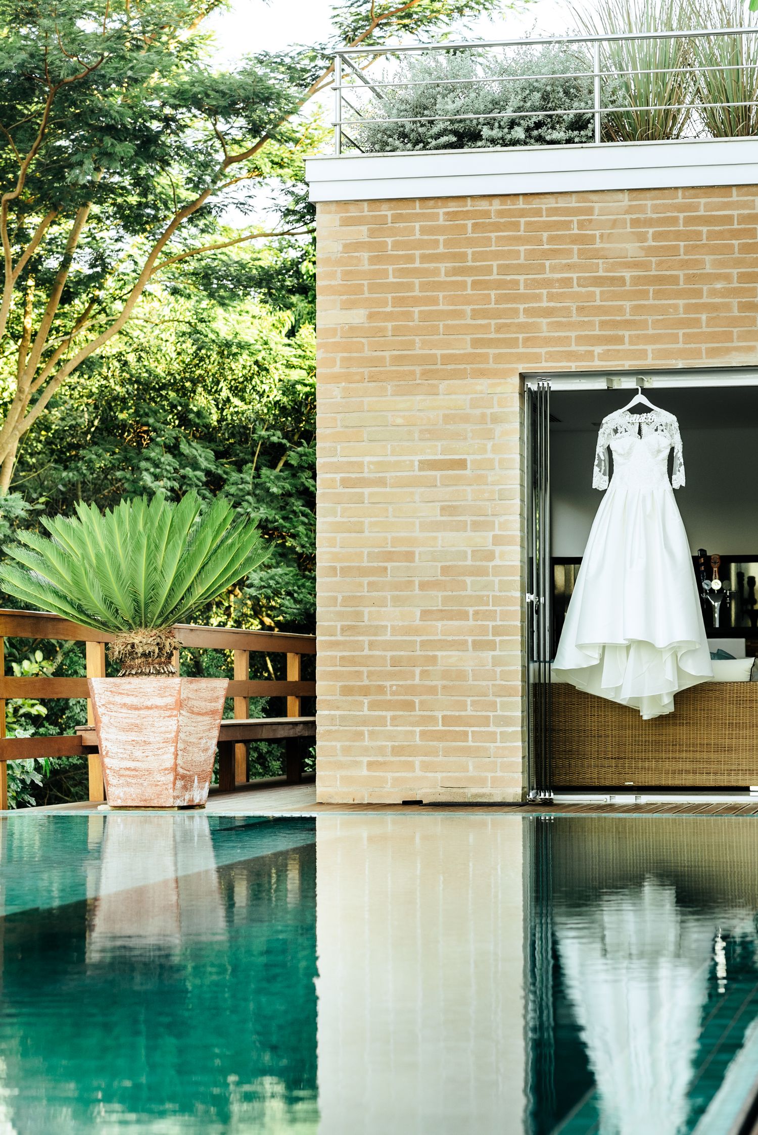White wedding dress hanging elegantly by turquoise pool with brick wall backdrop.