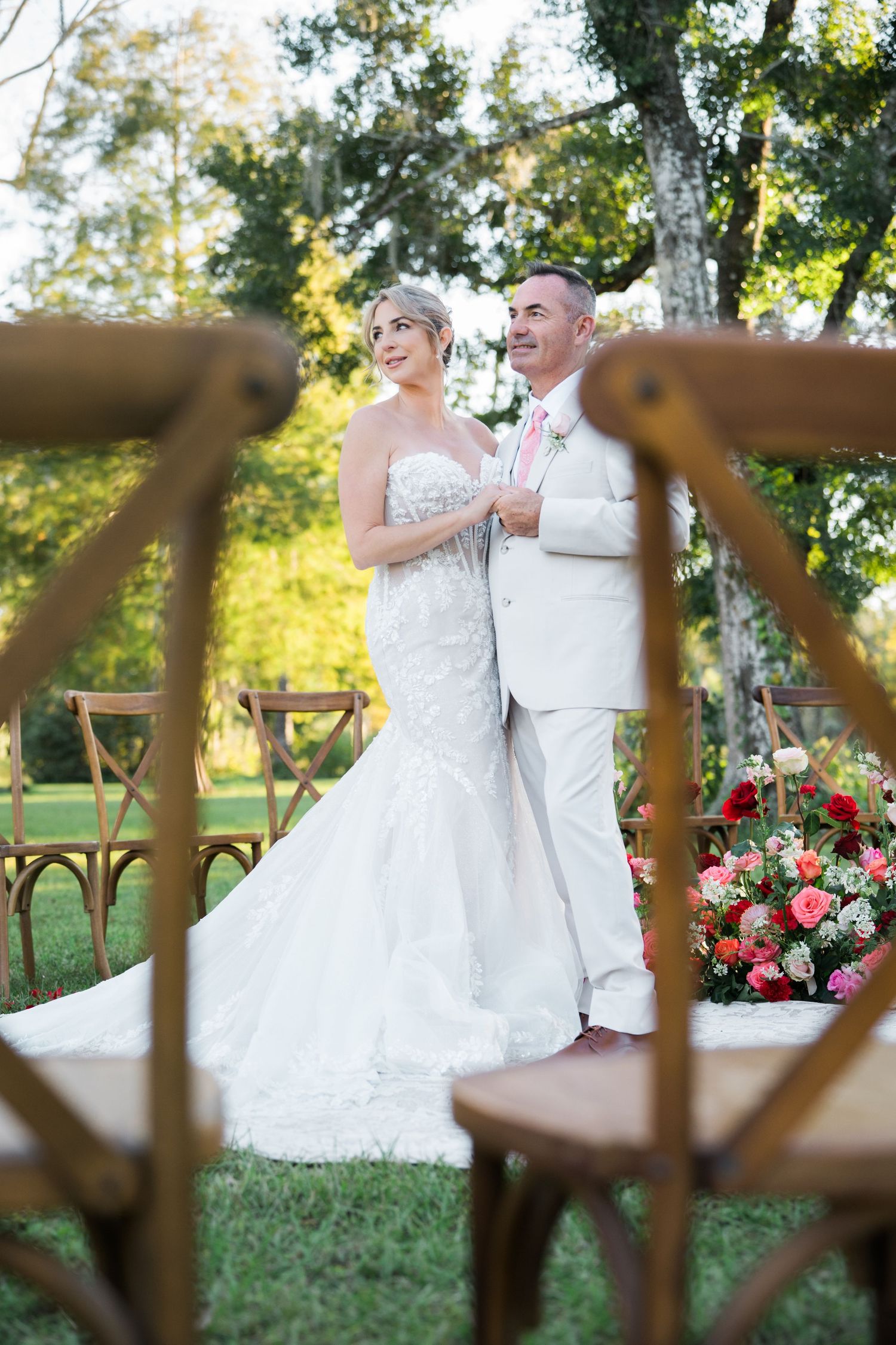Bride and groom pose together among wooden chairs in garden setting.