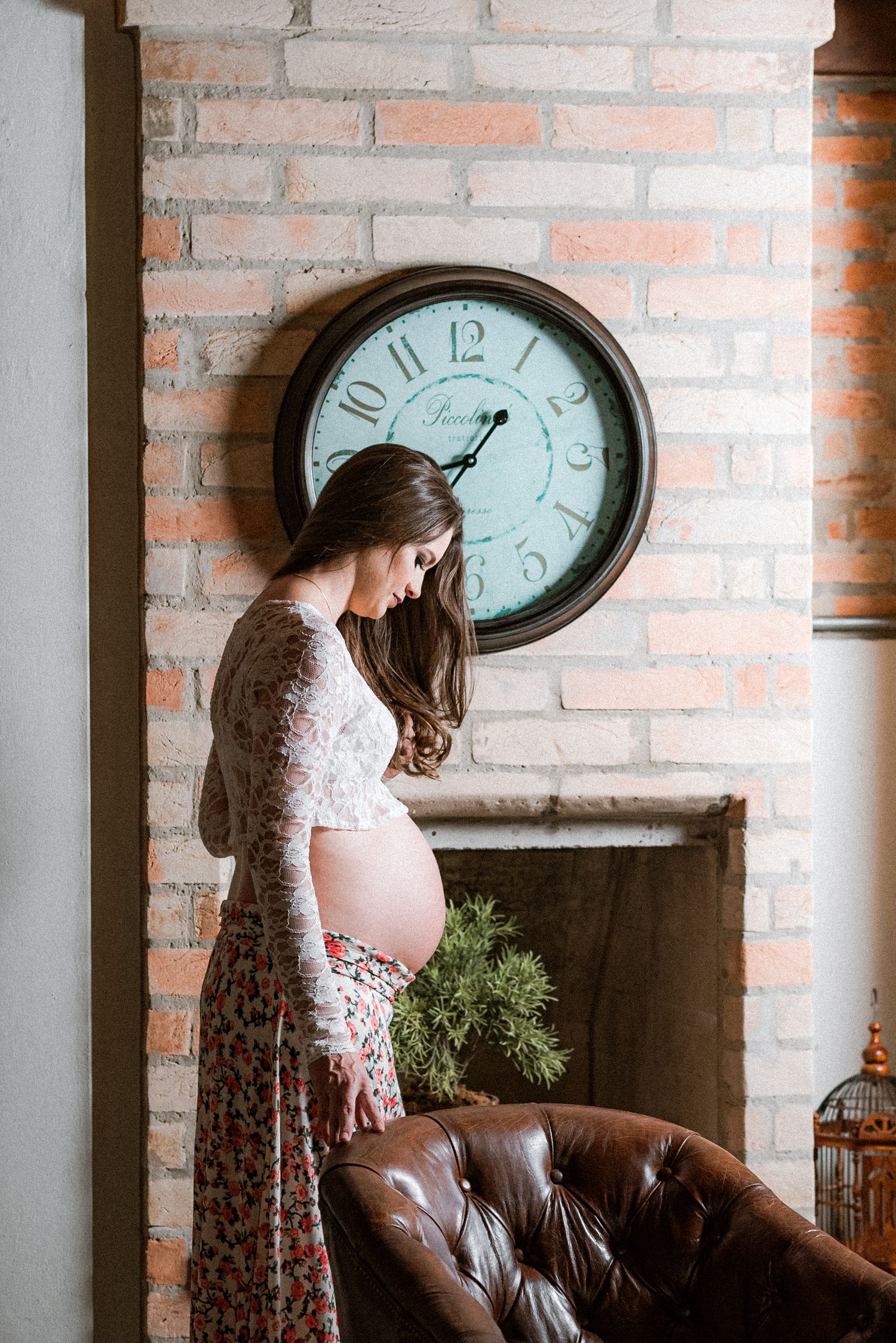 Person in floral dress poses near brick wall with vintage clock in rustic interior setting.
