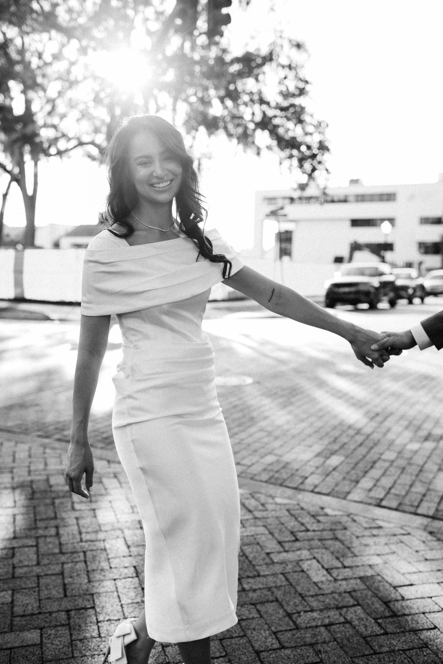 A bride in a white dress twirls joyfully on a brick pathway in sunlight.