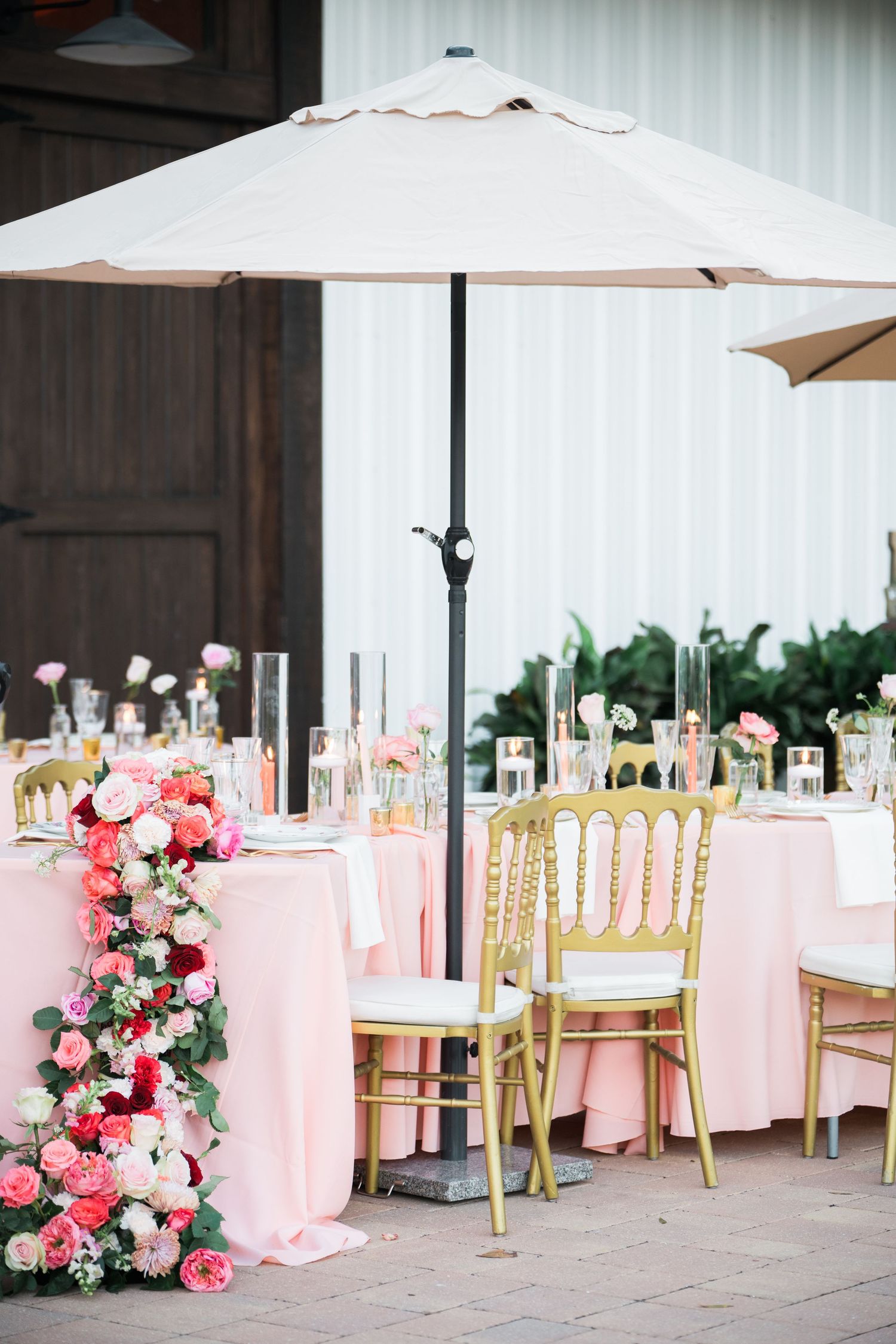Outdoor wedding reception setup with pink tablecloths and gold chiavari chairs under market umbrellas.