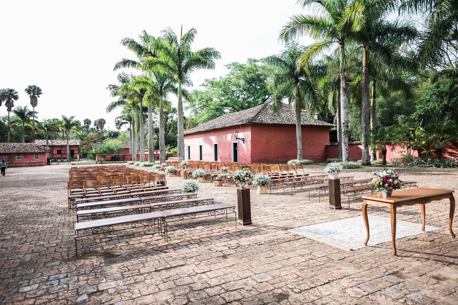 Outdoor wedding venue with wooden benches and pink buildings surrounded by palm trees.
