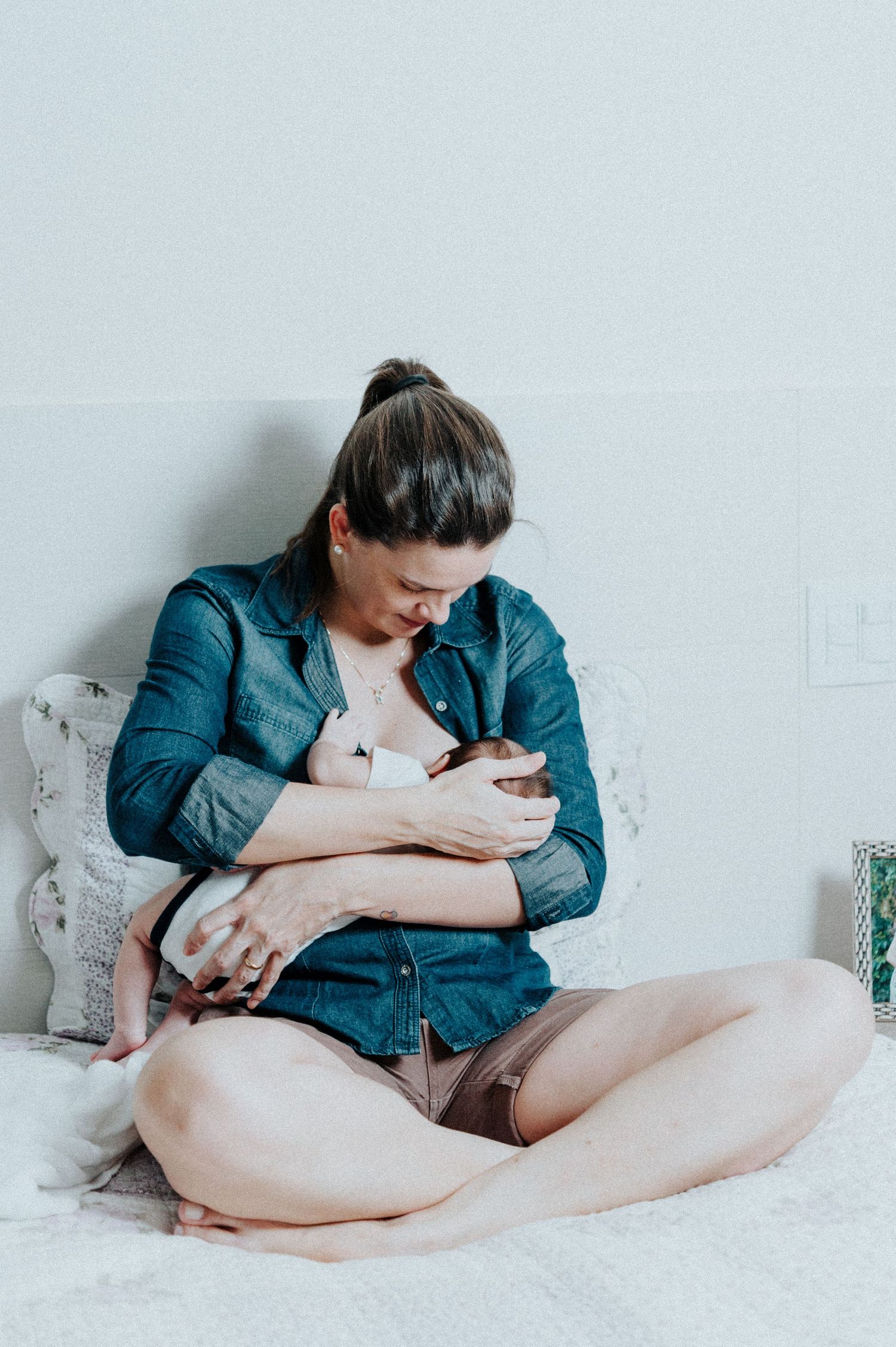 Mother in denim shirt nursing baby on white bedding in natural light.