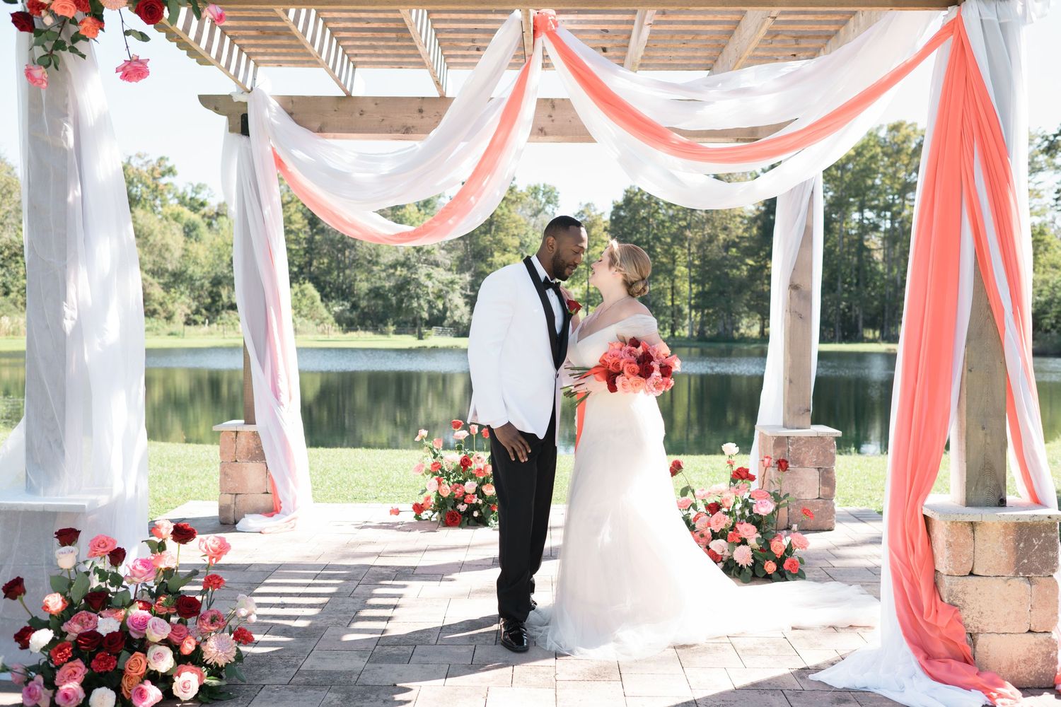 Wedding ceremony setup with coral and white draped gazebo by lakeside.