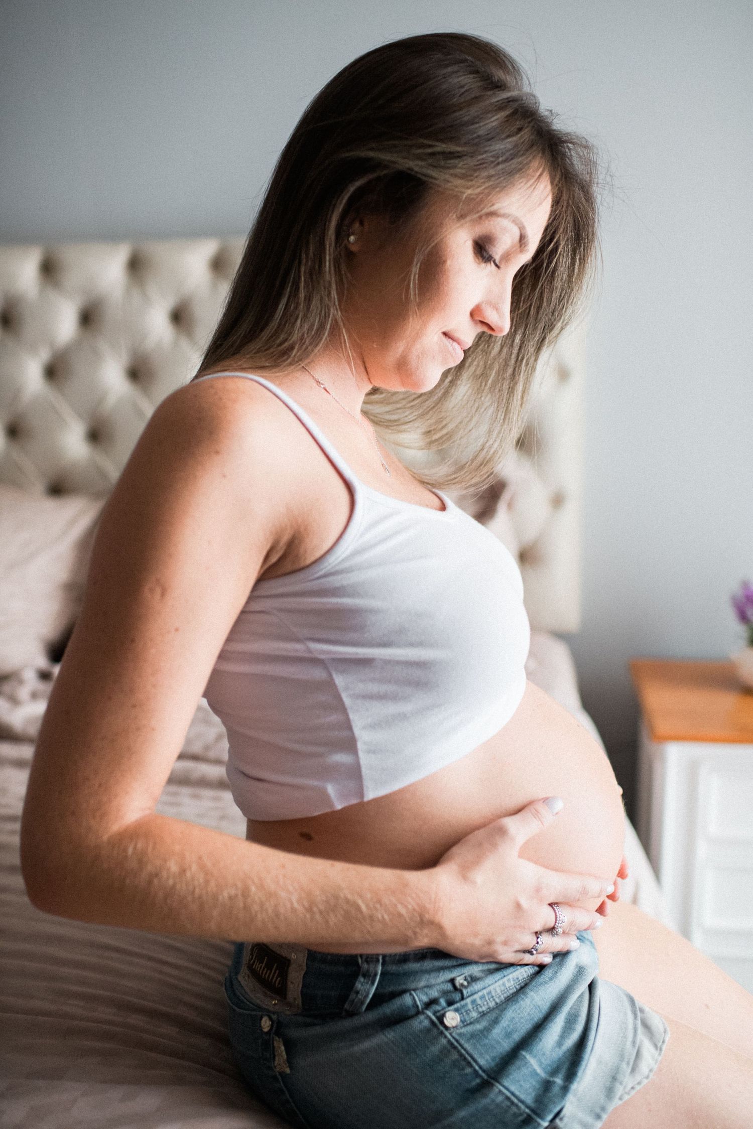 Maternity photos showing woman in white tank top and jeans on tufted headboard bed.